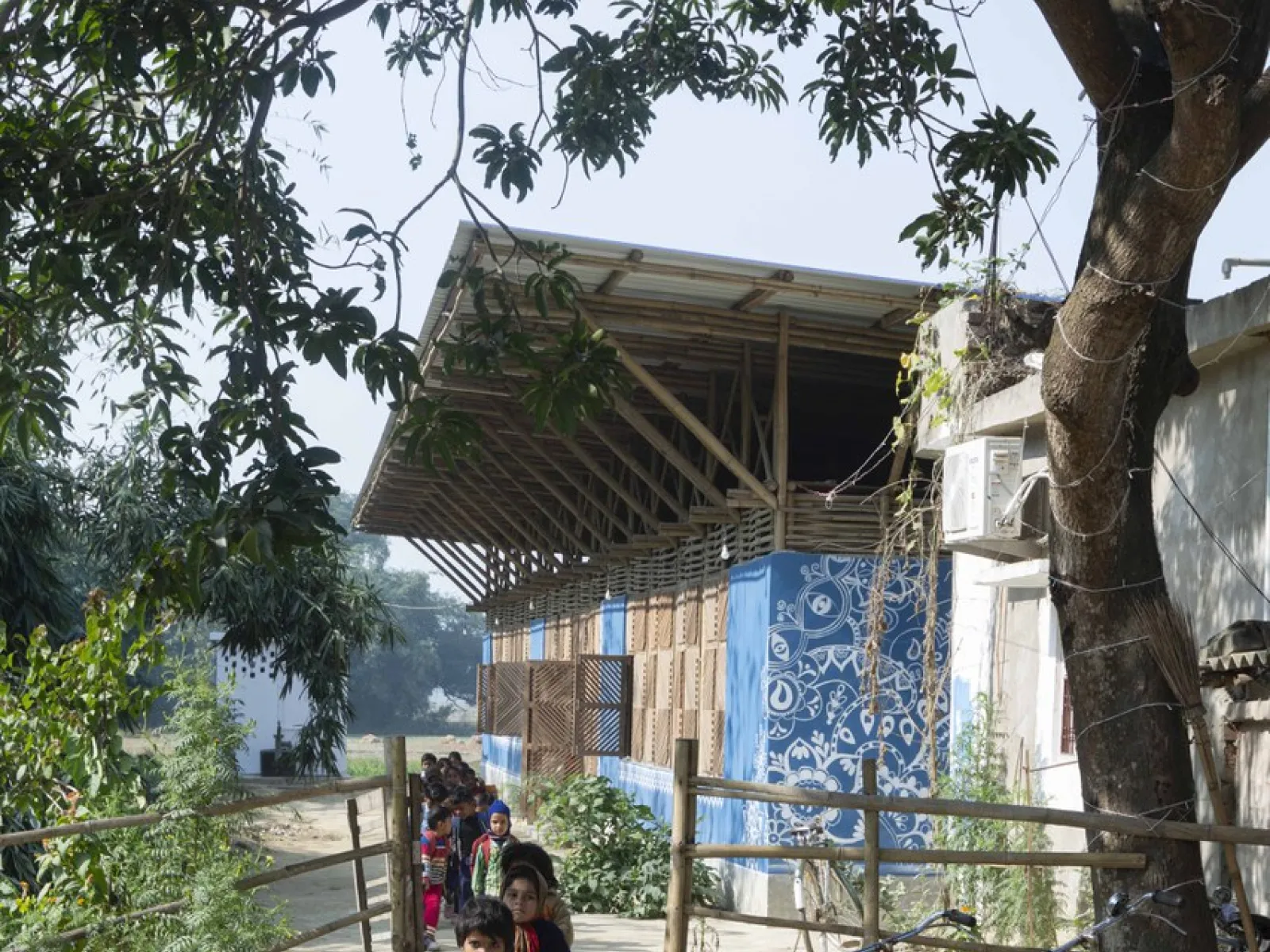 Eine Gruppe von Kindern geht einen Weg entlang, der zu einem nachhaltig gebauten Gebäude mit blauen Wänden und einem großen Dach führt. Im Vordergrund sind Bäume und ein Zaun zu sehen. Die Szene vermittelt eine ländliche, sonnige Atmosphäre.