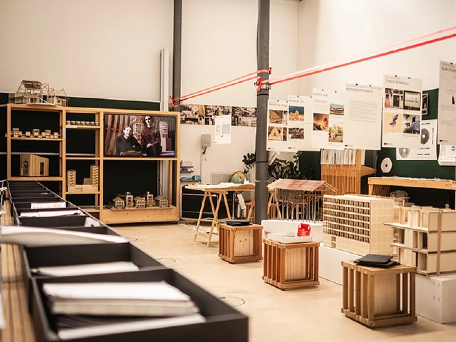 Ein Raum der Liechtenstein School of Architecture mit Ausstellungsstücken und Modellen. Im Vordergrund sind Tische mit Architekturmodellen, im Hintergrund ein Bildschirm mit einer Präsentation. An den Wänden hängen Plakate mit architektonischen Entwürfen.