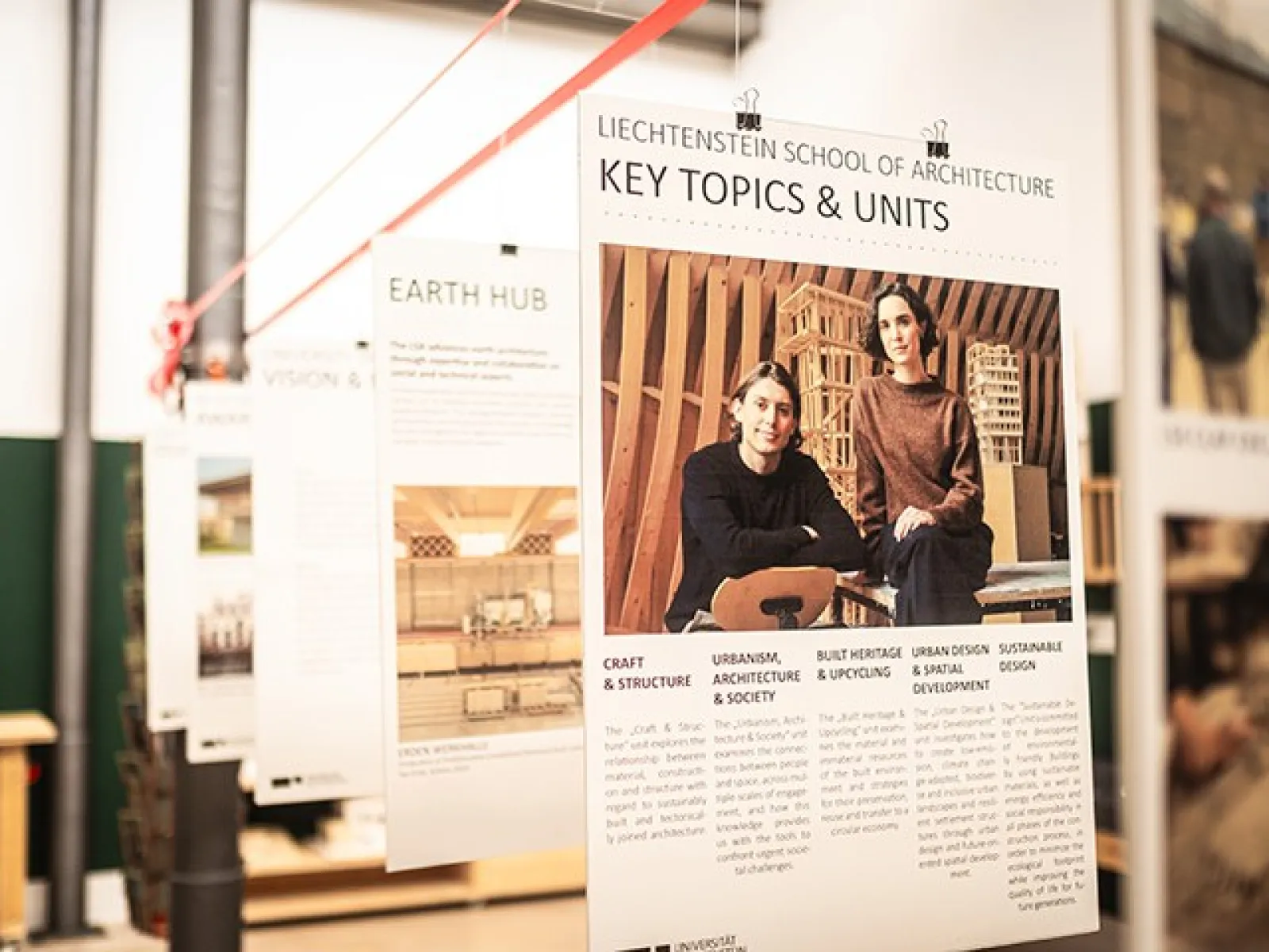 Ein Ausstellungsraum mit Architekturmodellen und Informationspostern über Lehmbau-Experten. Im Vordergrund ein detailliertes Modell eines Gebäudes mit einem roten Dach.