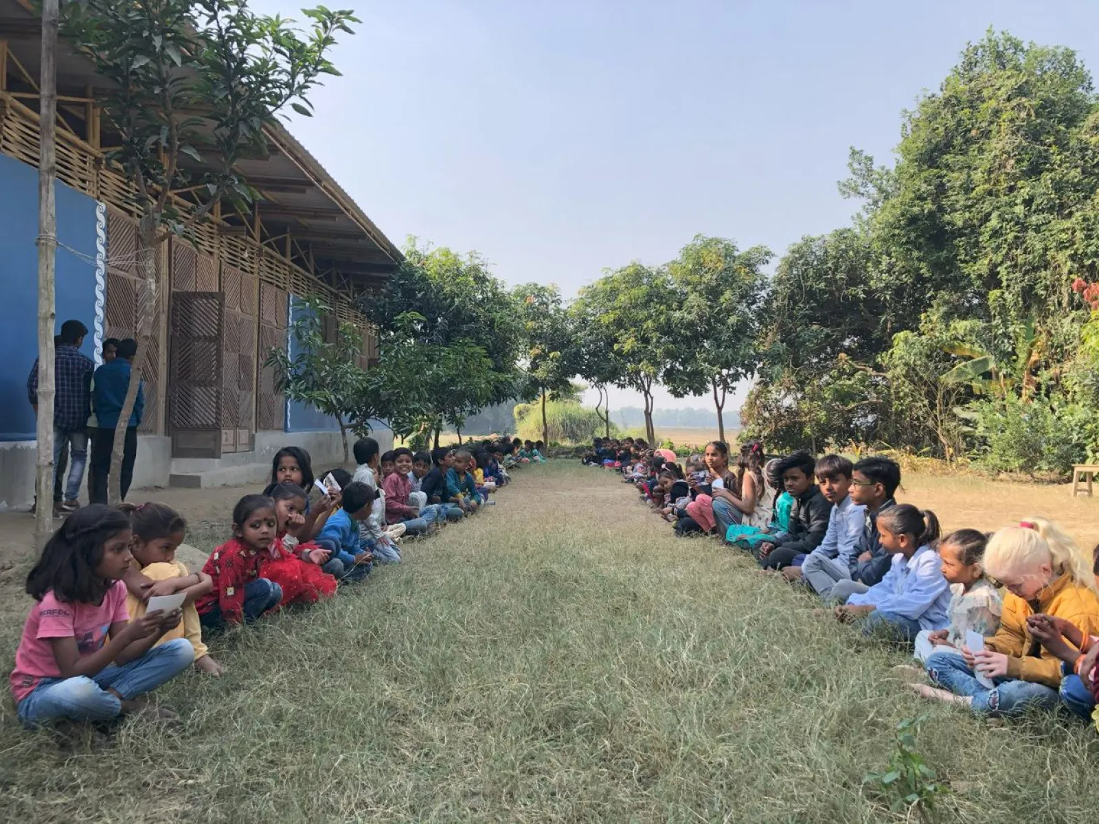 Kinder sitzen in zwei Reihen auf einer Wiese vor einem Schulgebäude in Indien. Sie sind von Bäumen umgeben und scheinen an einer Aktivität teilzunehmen. Die Szene vermittelt Gemeinschaft und Bildung im Freien.
