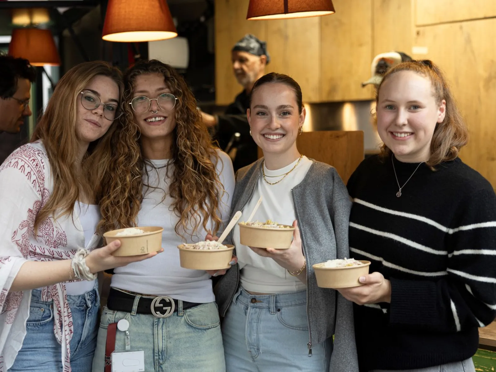 Vier lächelnde Personen halten Schalen mit Essen in einem gemütlichen Innenraum. Im Hintergrund ist eine Küche zu sehen. Die Szene vermittelt eine freundliche und einladende Atmosphäre.