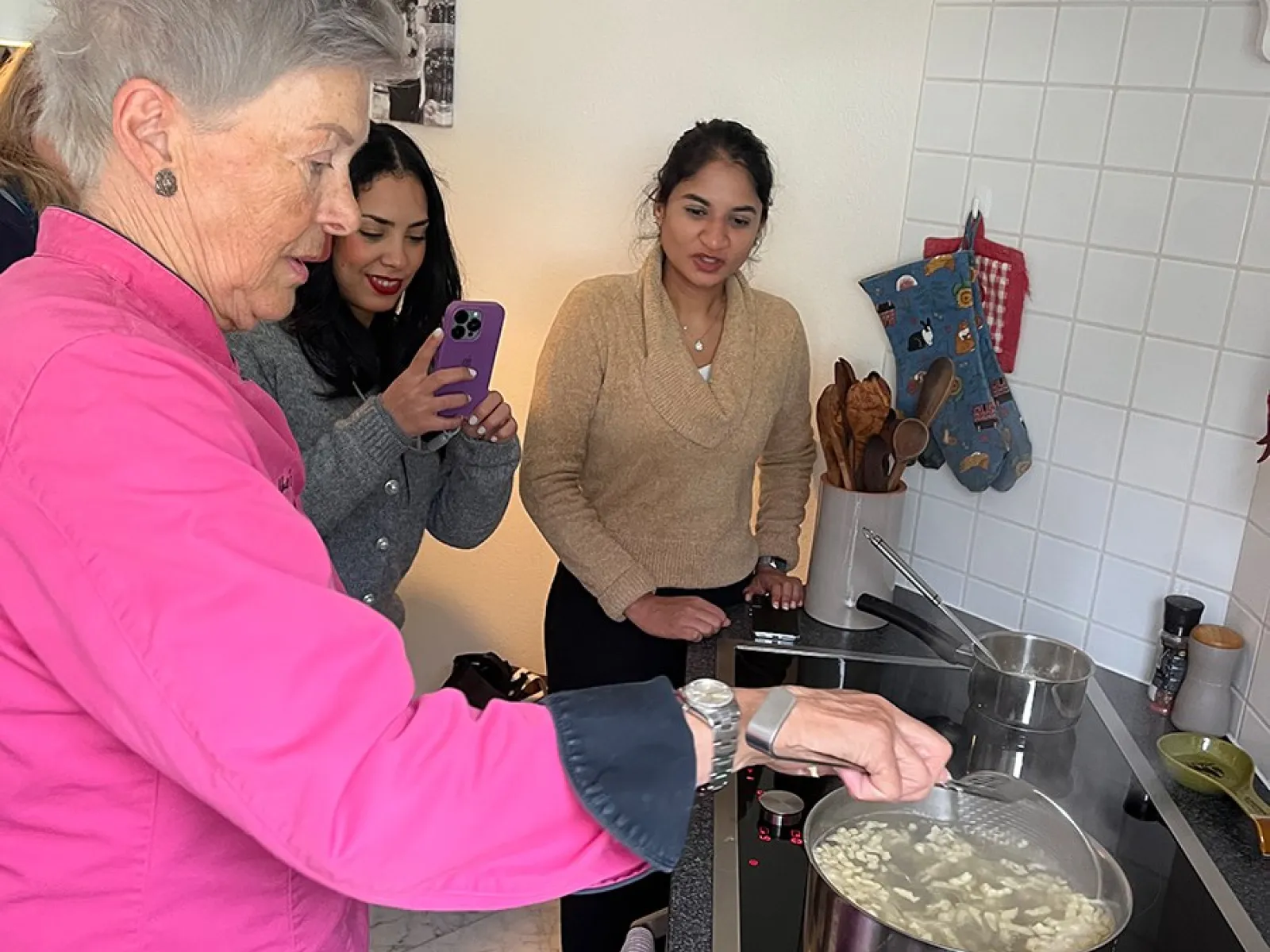 Eine Gruppe von Menschen steht in einer Küche. Eine Person in einem pinken Oberteil kocht an einem Herd, während zwei andere Personen zuschauen und eine von ihnen mit einem Smartphone fotografiert. Auf dem Herd steht ein Topf mit kochendem Wasser.