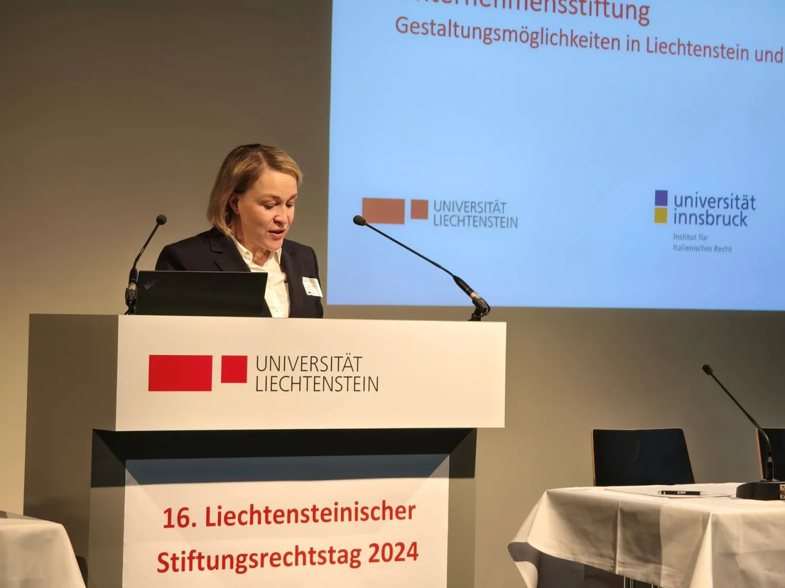 Eine Rednerin steht am Podium während des 16. Liechtensteinischen Stiftungsrechtstags 2024 an der Universität Liechtenstein. Im Hintergrund ist eine Präsentationsfolie mit Logos der Universität Liechtenstein und der Universität Innsbruck zu sehen.