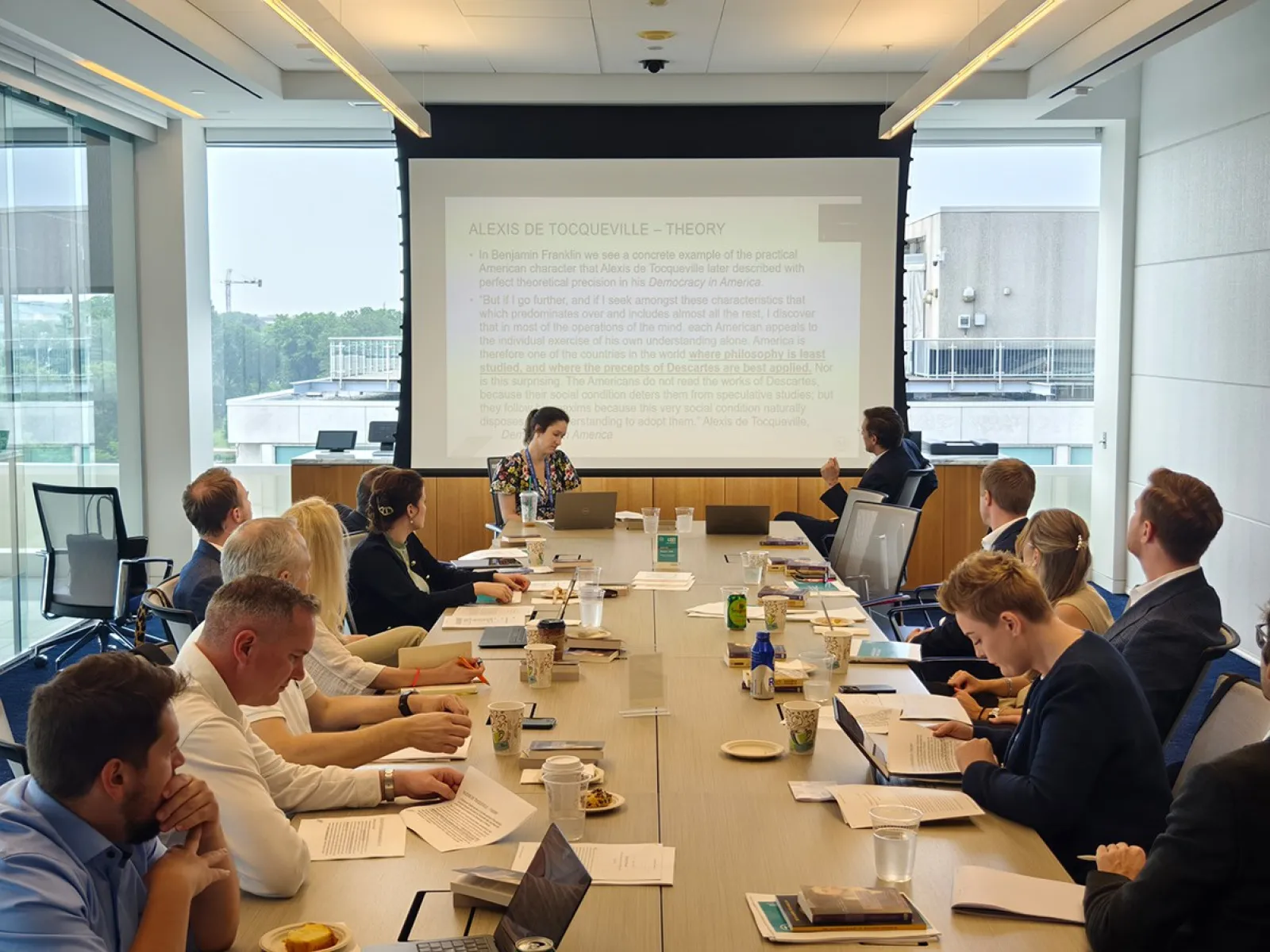 Ein Konferenzraum mit mehreren Personen, die an einem langen Tisch sitzen und einer Präsentation folgen. Im Hintergrund ist eine große Leinwand mit projiziertem Text zu sehen. Die Teilnehmer sind in eine Diskussion vertieft.