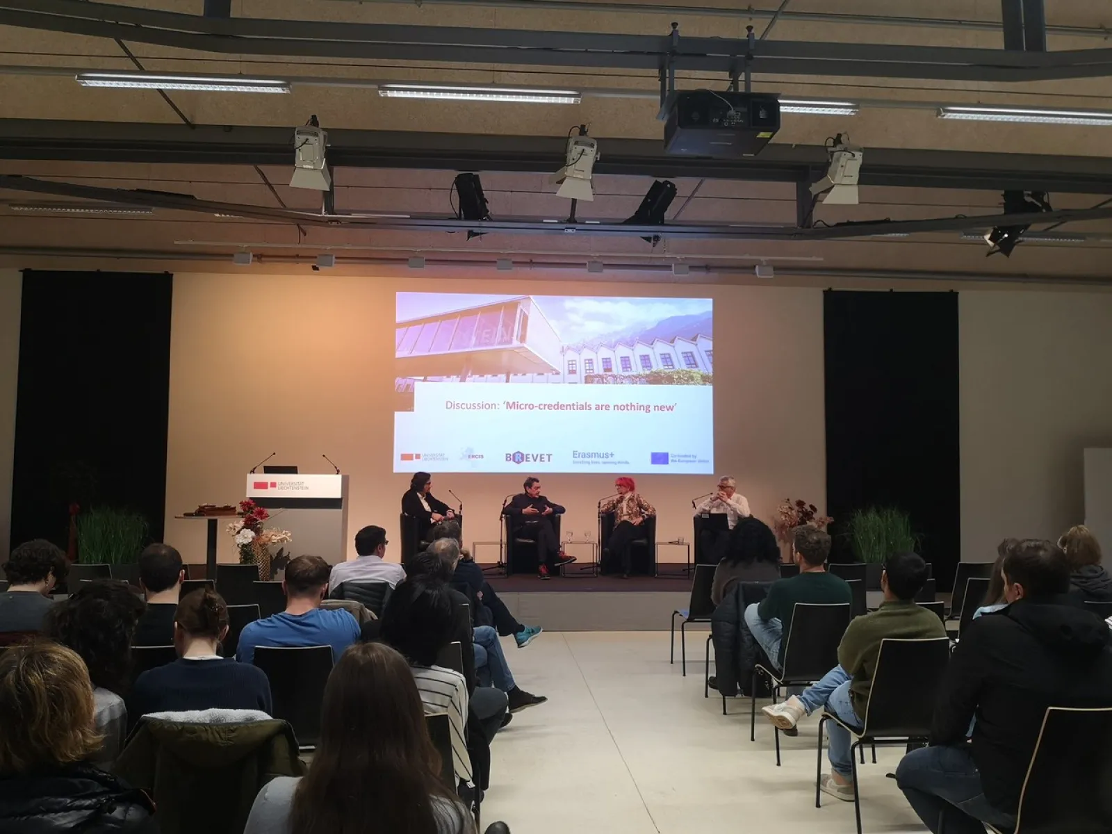Ein Auditorium mit mehreren Personen, die einer Podiumsdiskussion zuhören. Drei Redner sitzen auf der Bühne vor einer Leinwand mit der Aufschrift „Discussion: 'Micro-credentials are nothing new'“. Die Veranstaltung findet in einem modernen Konferenzraum statt.