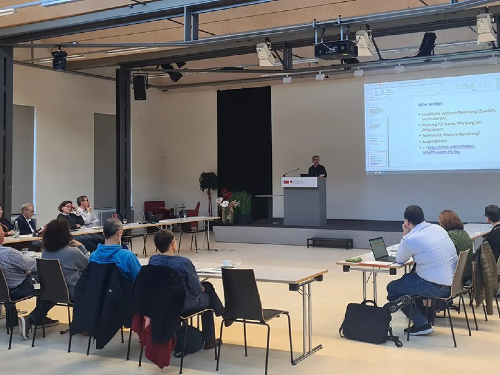 Eine Gruppe von etwa 20 Personen sitzt in einem Auditorium der Universität Liechtenstein, während ein Vortragender an einem Rednerpult spricht. Auf einer großen Leinwand ist eine Präsentation mit dem Titel "Wie weiter" zu sehen.