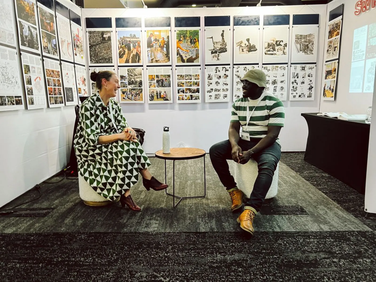 Zwei Personen sitzen in einer Ausstellung vor einer Wand mit zahlreichen Bildern und Texten. Sie unterhalten sich entspannt auf Hockern. Die Ausstellung thematisiert urbanes Design und soziale Realitäten in Afrika.
