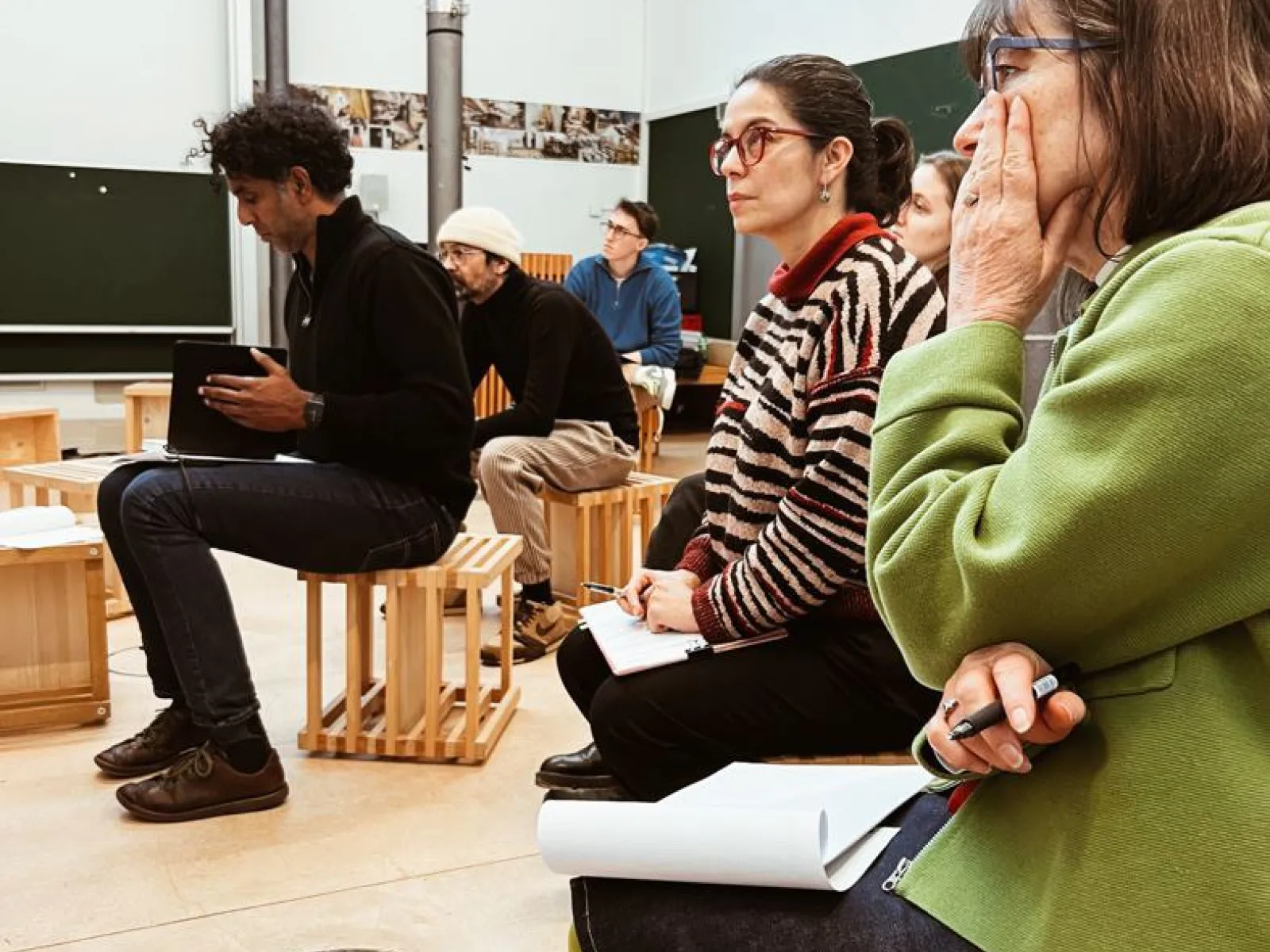 Eine Gruppe von Personen sitzt aufmerksam in einem Seminarraum. Sie sind auf Holzhockern platziert und scheinen einer Präsentation oder Diskussion zu folgen. Im Hintergrund sind Tafeln und Poster zu sehen. Die Szene vermittelt eine konzentrierte Lernatmosphäre.