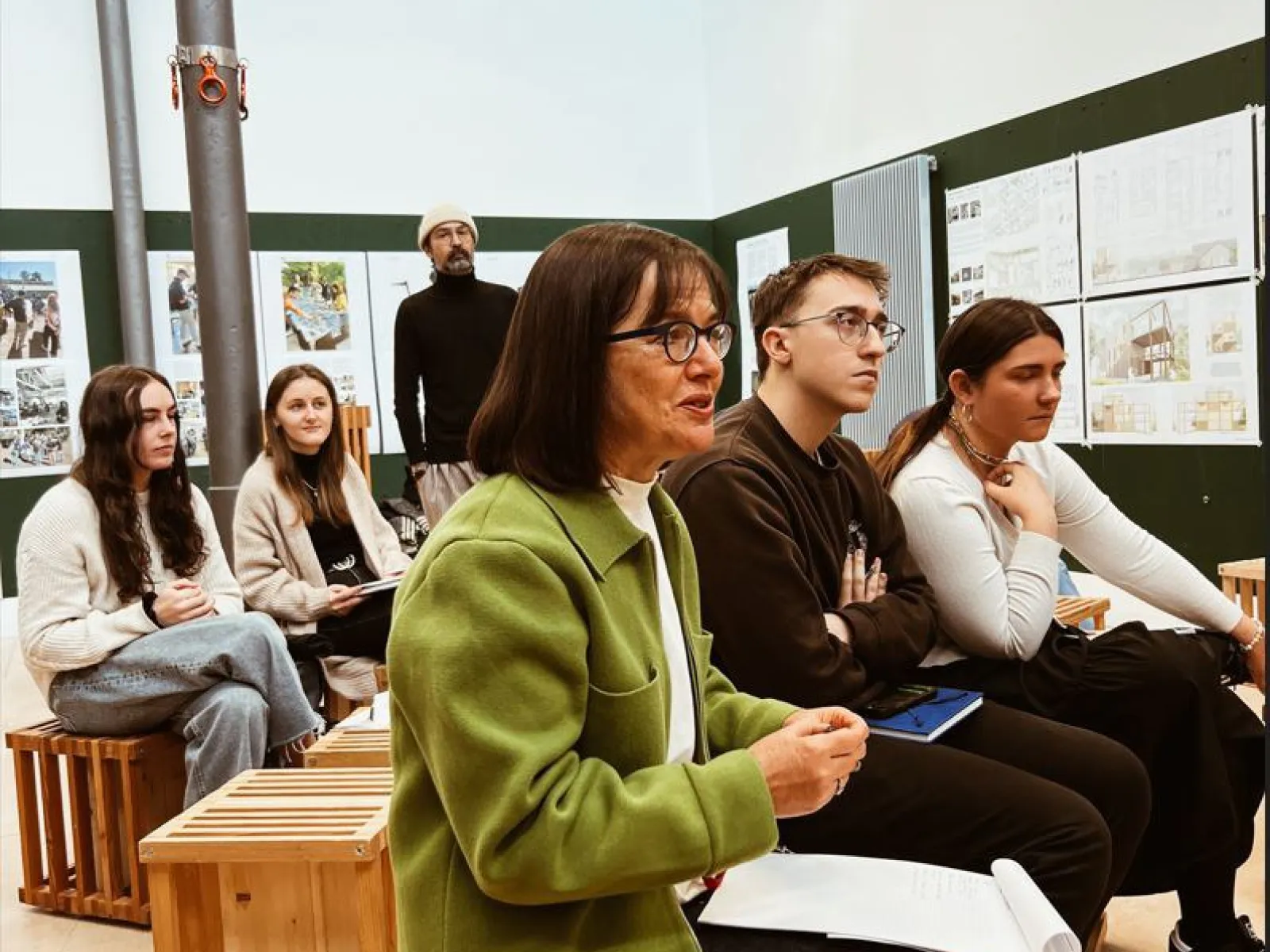 Eine Gruppe von Studierenden und Dozenten sitzt in einem Seminarraum und hört aufmerksam zu. Im Hintergrund sind Präsentationstafeln mit städtischen Entwürfen zu sehen.
