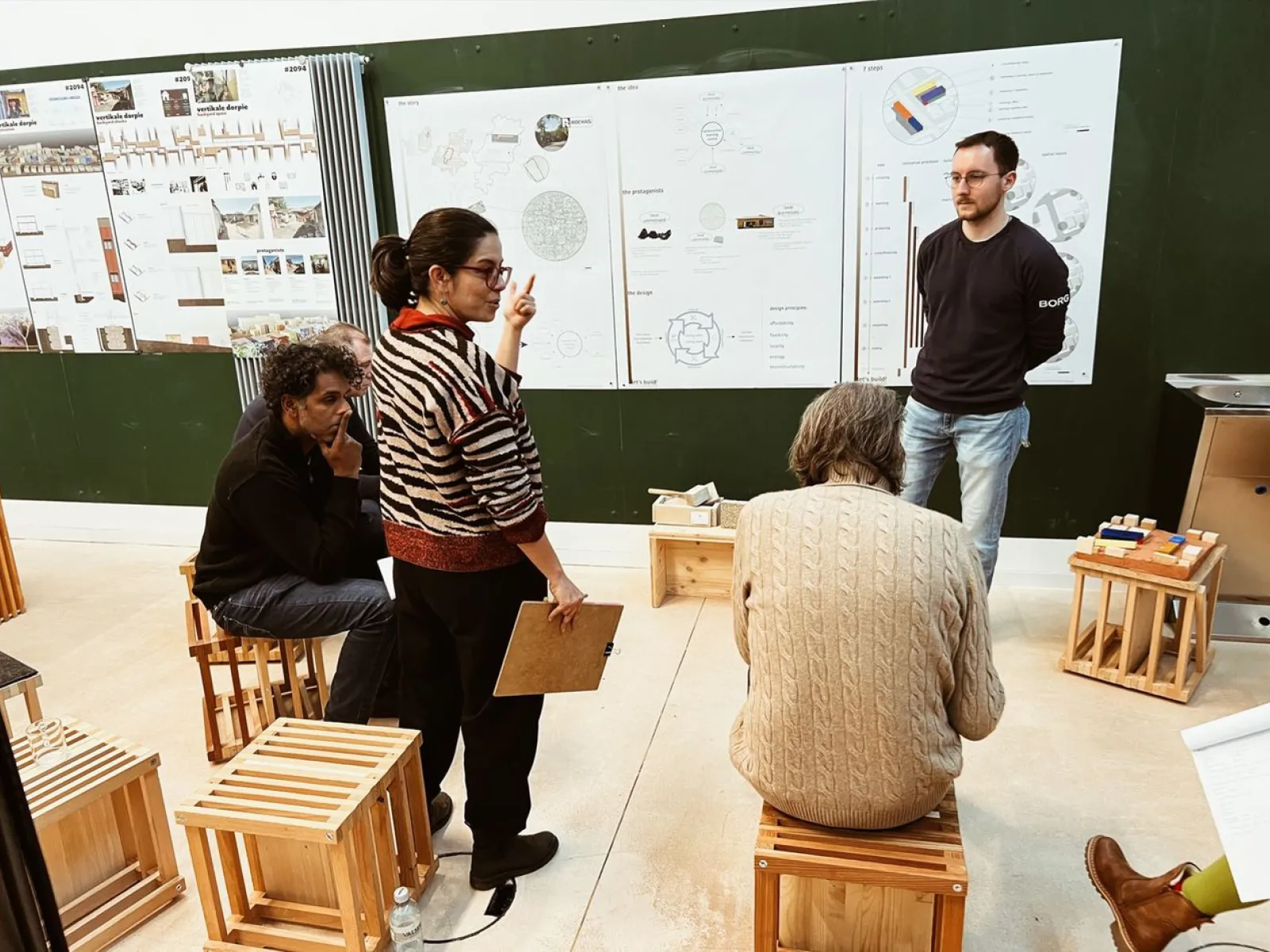 Eine Gruppe von Personen diskutiert vor einer Wand mit Architekturplänen. Sie sitzen auf Holzhockern in einem Seminarraum. Eine Frau steht und gestikuliert, während sie auf die Pläne zeigt.