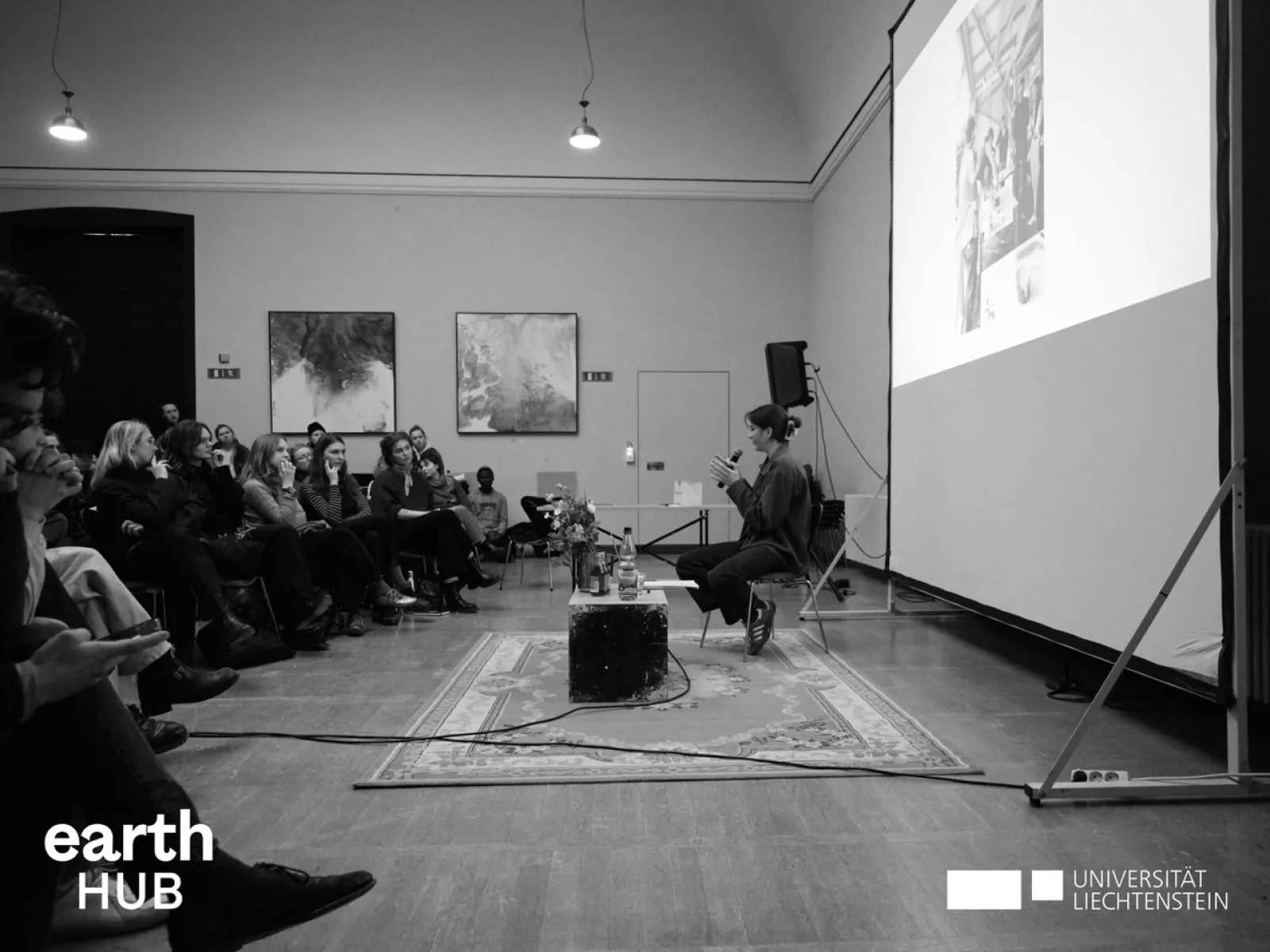 Schwarz-Weiß-Foto eines Vortrags an der Universität Liechtenstein. Eine Person sitzt vor einem Publikum und spricht, während eine Präsentation auf eine Leinwand projiziert wird. Im Vordergrund sind Logos von "earth HUB" und "Universität Liechtenstein" sichtbar.