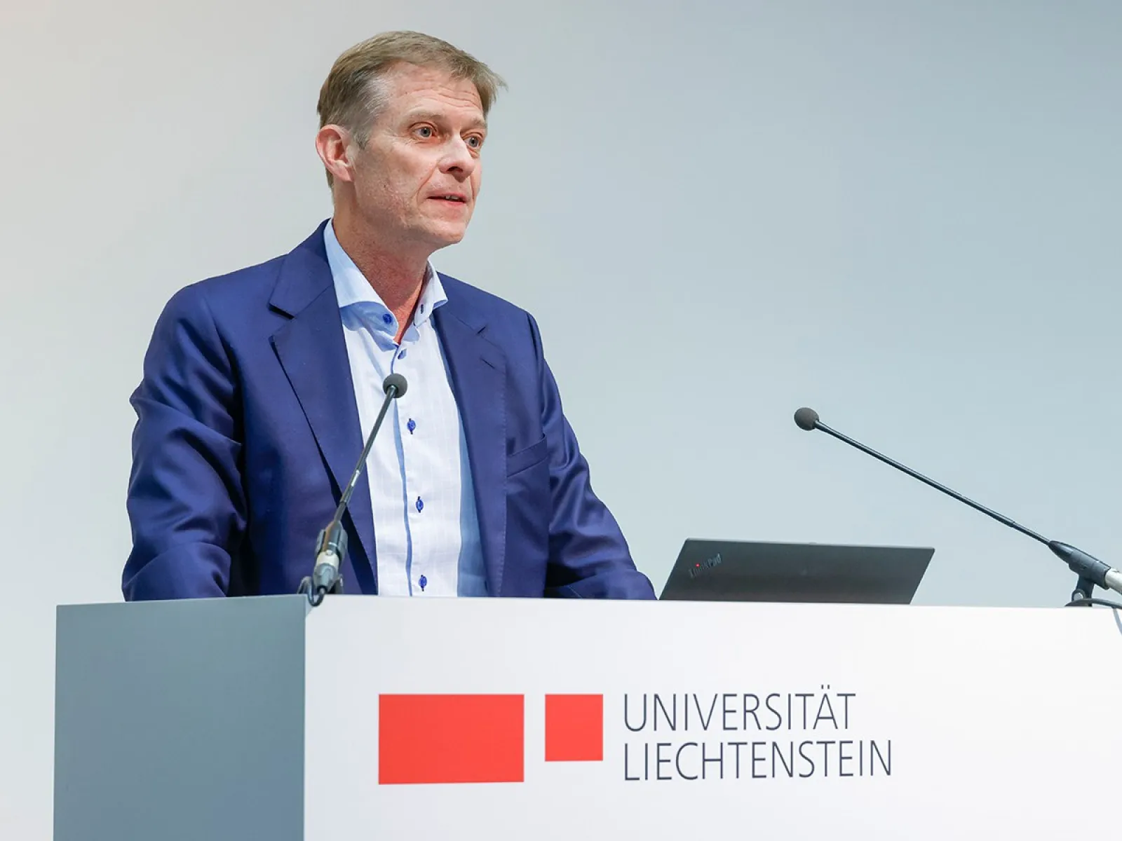 Ein Mann steht an einem Rednerpult mit dem Logo der Universität Liechtenstein und spricht in ein Mikrofon.