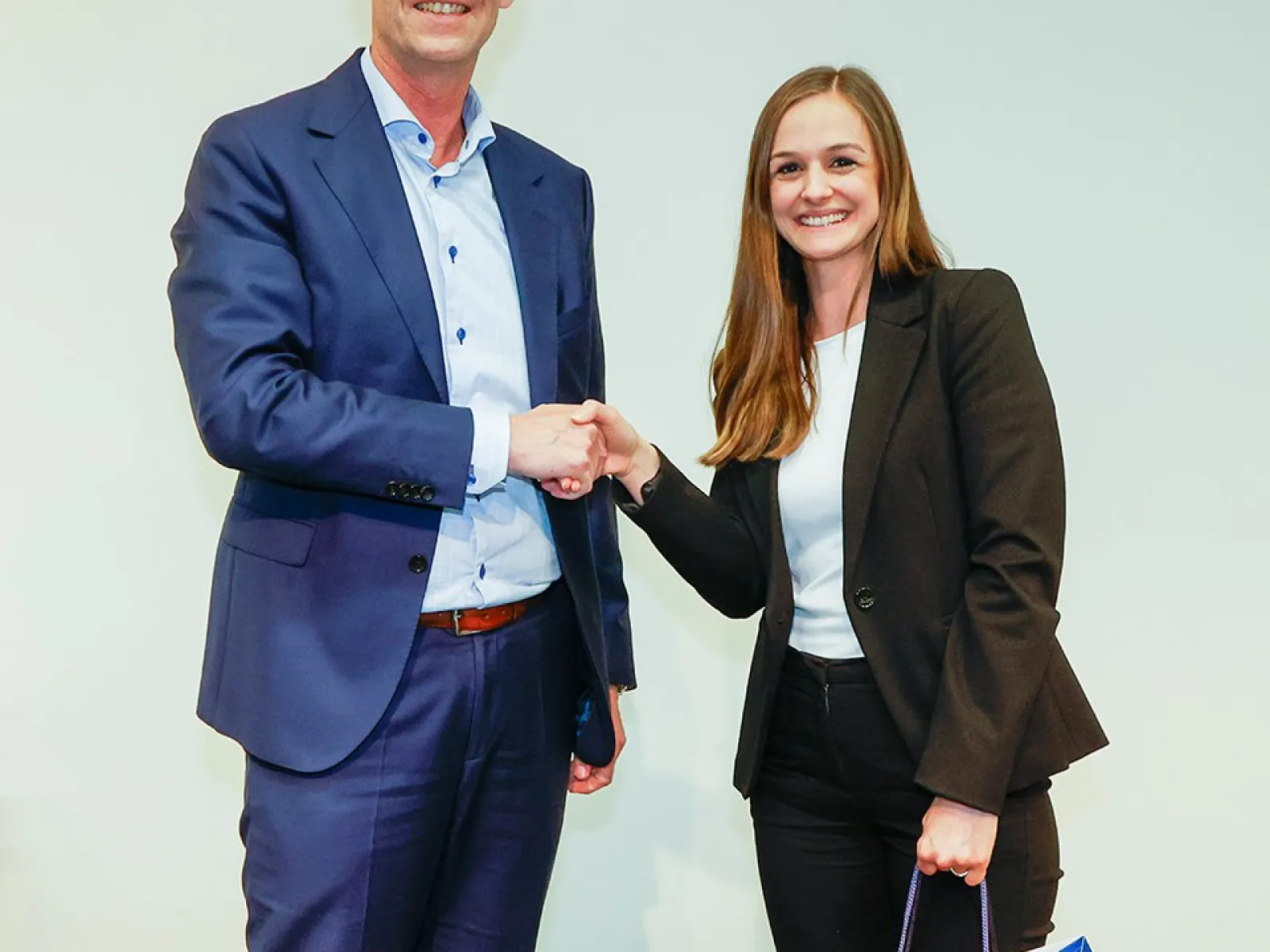 Zwei Personen stehen auf einer Bühne und schütteln sich die Hand. Die Frau hält eine Geschenktasche mit dem Logo der Universität Liechtenstein. Beide lächeln und tragen formelle Kleidung.