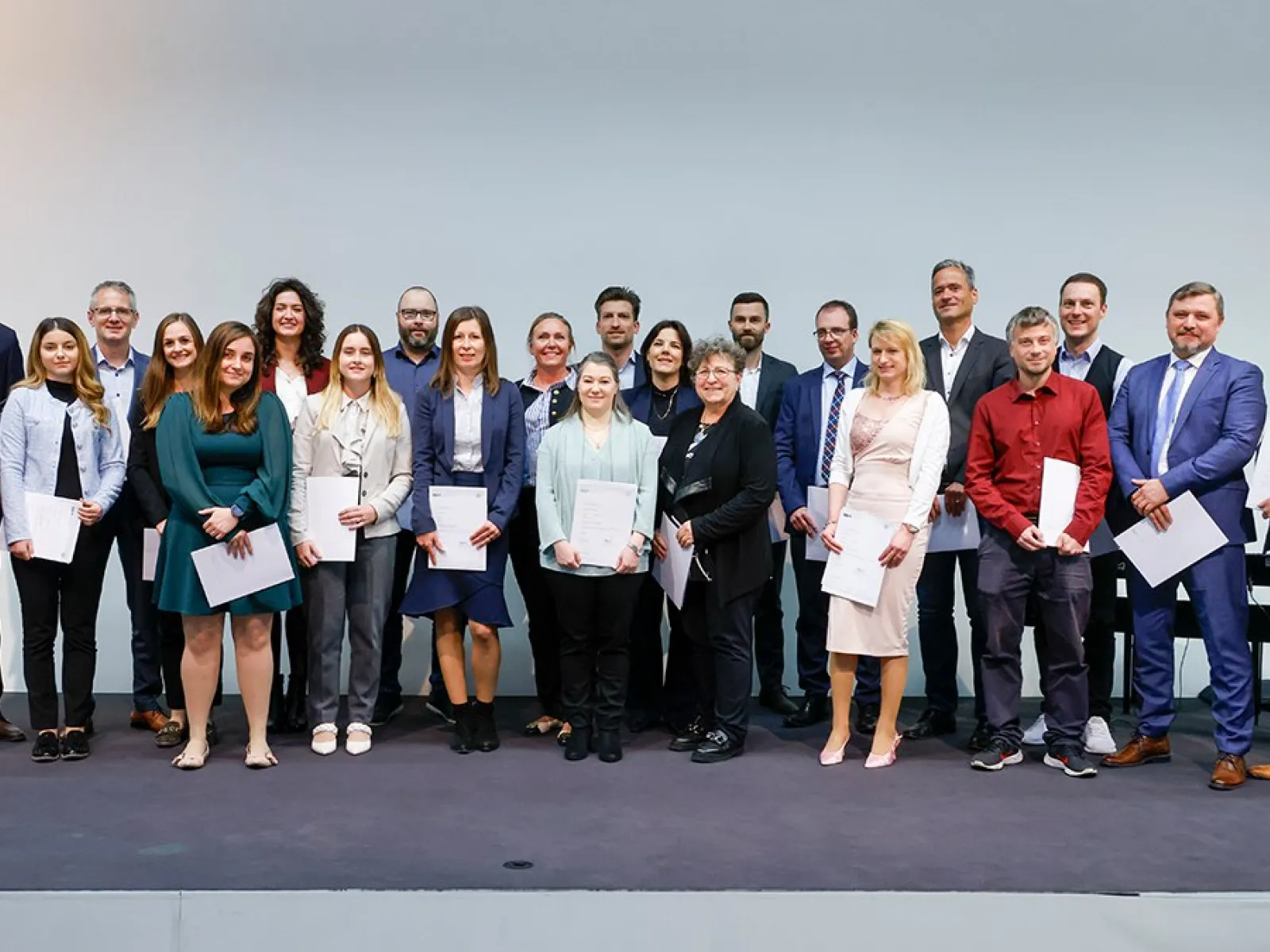 Eine Gruppe von Absolventinnen und Absolventen steht auf einer Bühne und hält Zertifikate in den Händen. Sie lächeln in die Kamera. Die Personen sind formell gekleidet, und im Hintergrund ist eine neutrale Wand zu sehen.