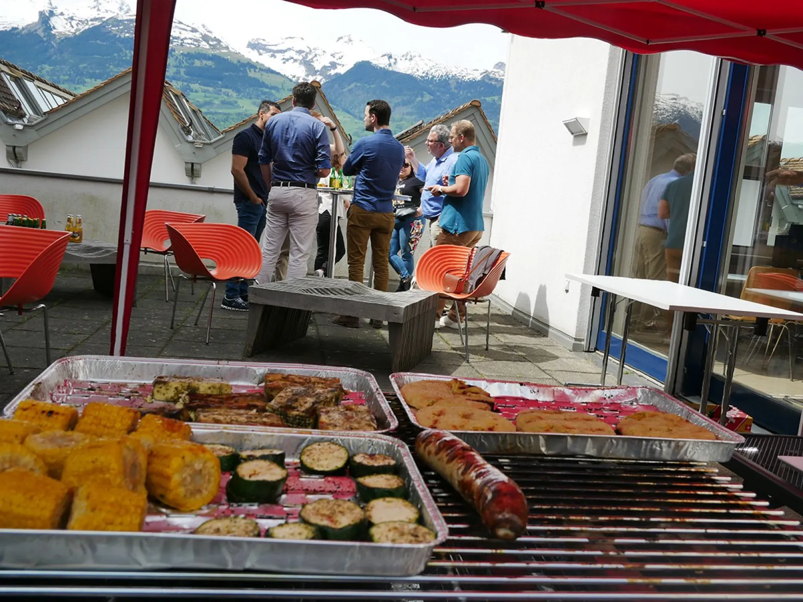 Alumni-Treffen an der Universität Liechtenstein: Eine Gruppe von Menschen steht auf einer Terrasse und unterhält sich bei einem BBQ. Im Vordergrund sind gegrillte Speisen zu sehen. Im Hintergrund sind Berge unter einem blauen Himmel sichtbar.
