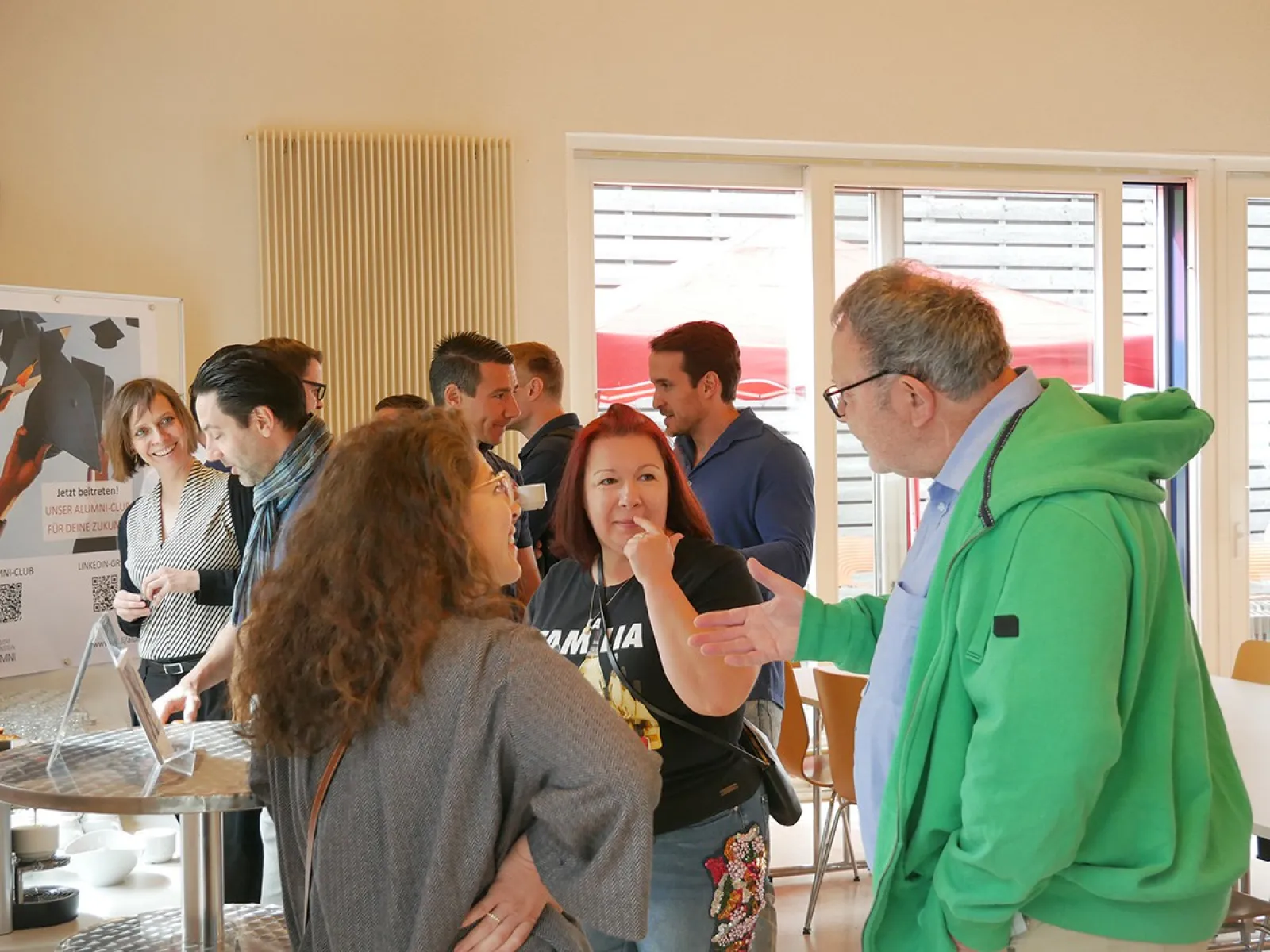 Eine Gruppe von Menschen unterhält sich lebhaft in einem hellen Raum auf dem Campus der Universität Liechtenstein. Im Hintergrund sind Fenster und ein Plakat zu sehen.