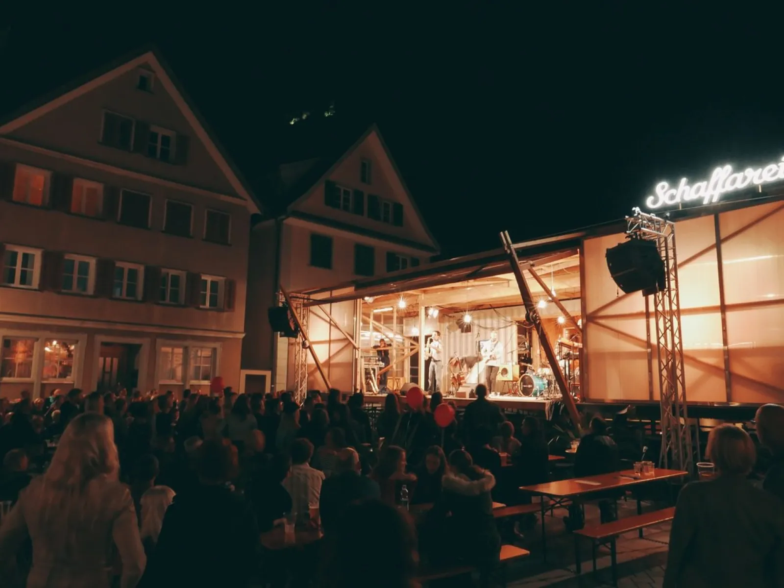 Eine beleuchtete Bühne des Base Camps der Universität Liechtenstein bei Nacht, umgeben von einem Publikum. Die Bühne ist modular und mobil, passend zur temporären Nutzung für Veranstaltungen und Stadtentwicklungsprojekte.