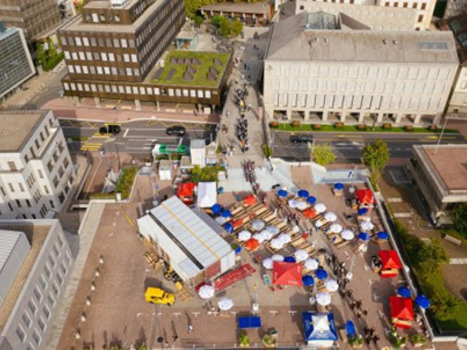 Luftaufnahme eines städtischen Platzes mit dem Base Camp der Universität Liechtenstein. Zu sehen sind modulare Strukturen, bunte Zelte und Menschen, die sich auf dem Platz bewegen. Gebäude umgeben den Platz, und eine Straße verläuft im Hintergrund.