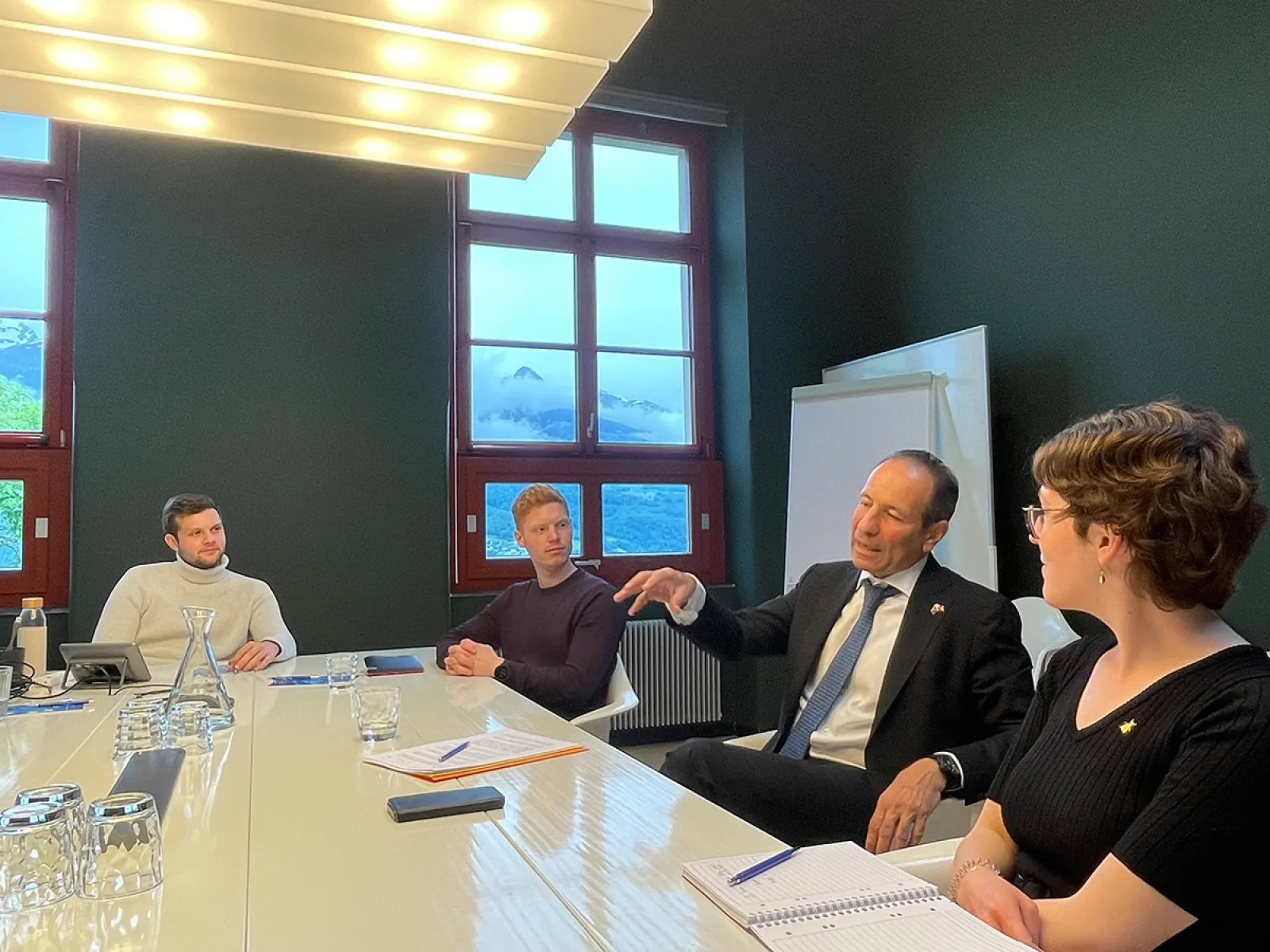 Ein Mann im Anzug spricht mit drei Personen an einem Konferenztisch in einem Raum mit großen Fenstern. Auf dem Tisch liegen Notizbücher und Gläser.