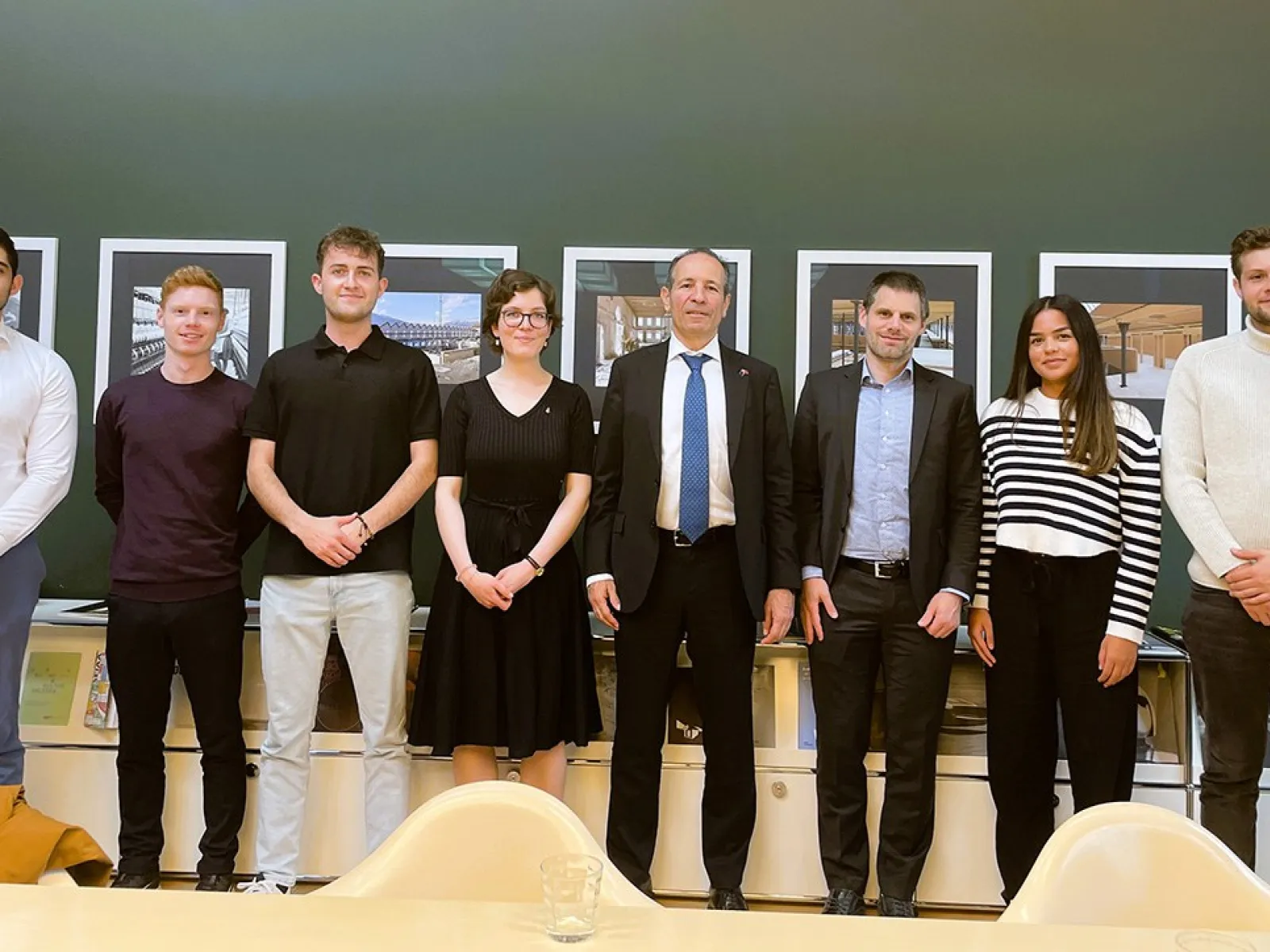 Eine Gruppe von sieben Personen, darunter Studierende und zwei Männer in Anzügen, steht lächelnd vor einer Wand mit Bildern. Sie befinden sich in einem Raum der Universität Liechtenstein.