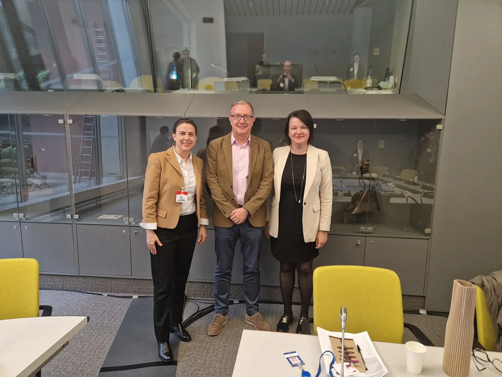 Drei Personen stehen in einem modernen Konferenzraum des Europarats in Straßburg. Sie lächeln in die Kamera. Im Hintergrund sind weitere Personen und Konferenztische zu sehen.