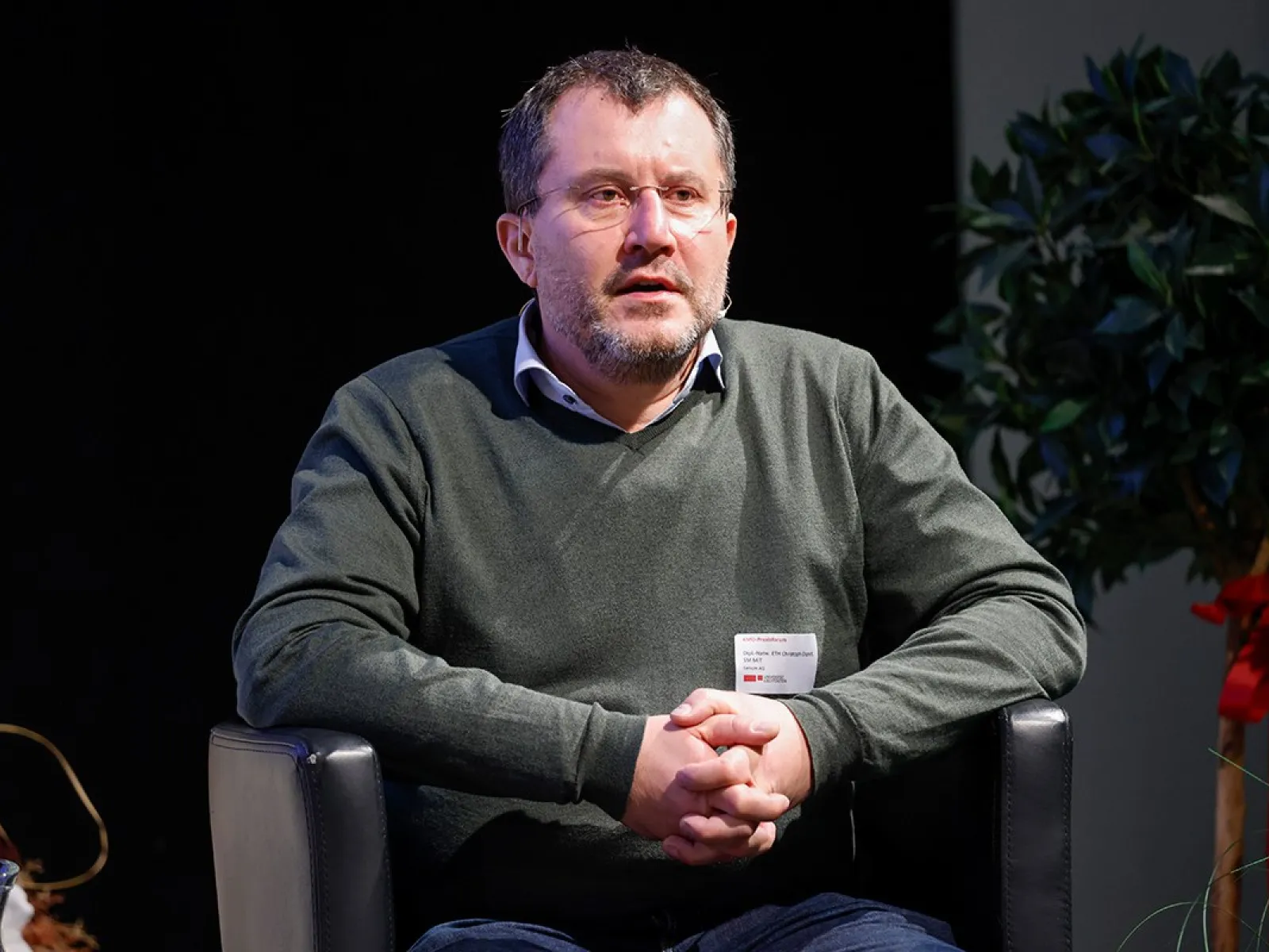 Eine Person sitzt auf einem schwarzen Sessel und hält die Hände gefaltet. Im Hintergrund sind Pflanzen zu sehen.