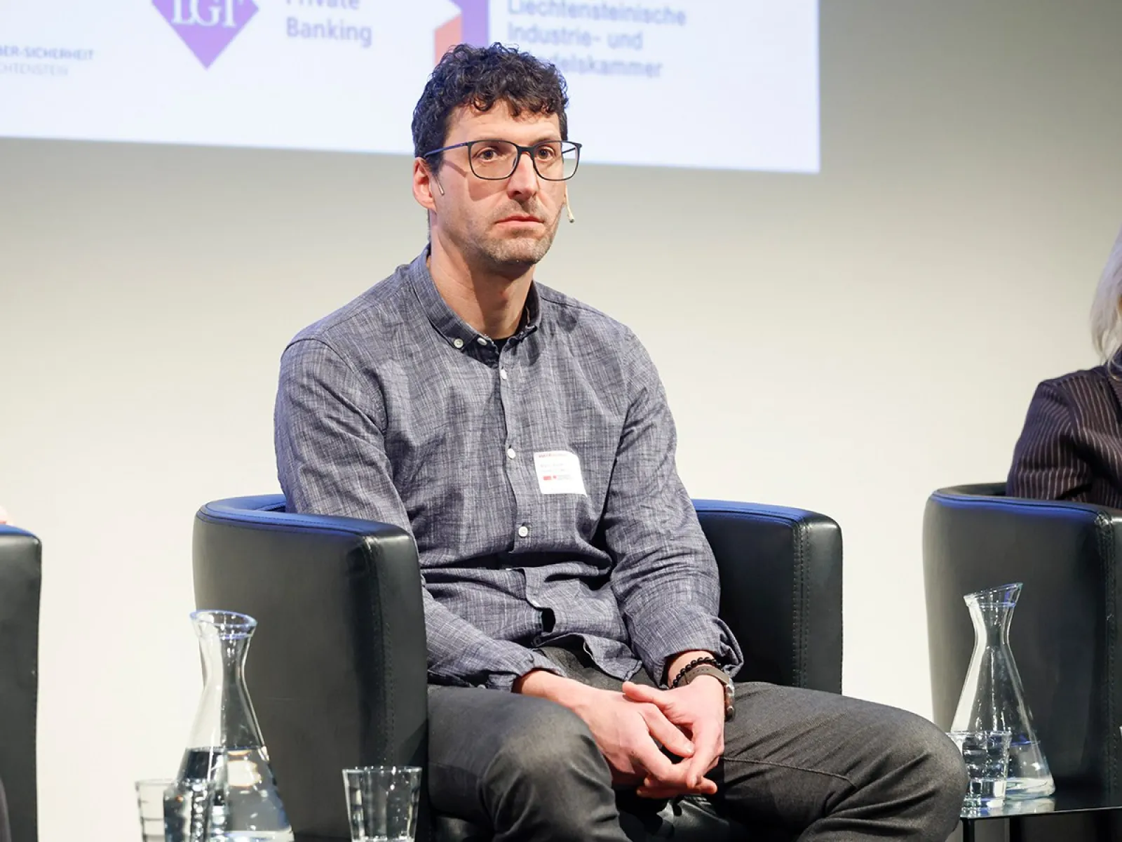 Eine Person sitzt auf einer Bühne in einem schwarzen Sessel. Im Hintergrund sind Logos von Sponsoren oder Partnern zu sehen. Auf dem Tisch vor der Person stehen Wasserkaraffen und Gläser.