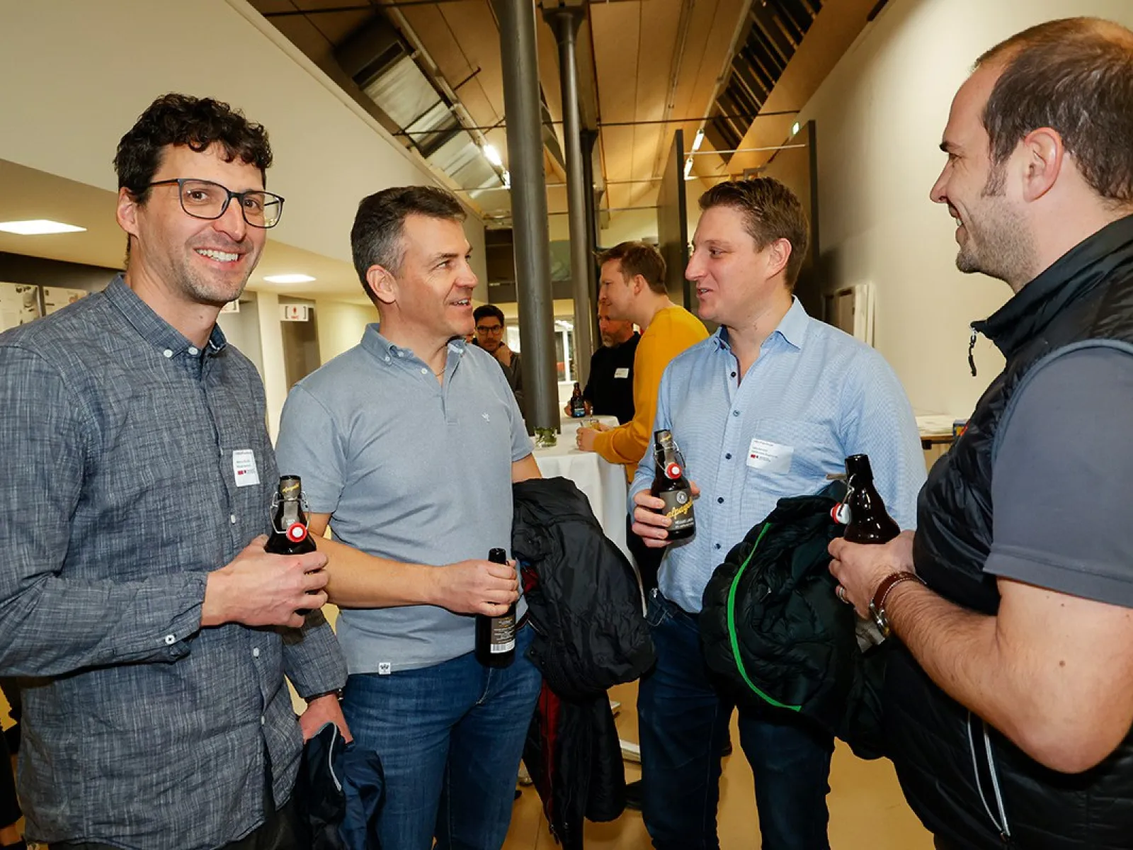 Vier Männer unterhalten sich bei einer Veranstaltung an der Universität Liechtenstein. Sie halten Getränke in der Hand und tragen Namensschilder. Die Atmosphäre wirkt entspannt und gesellig.