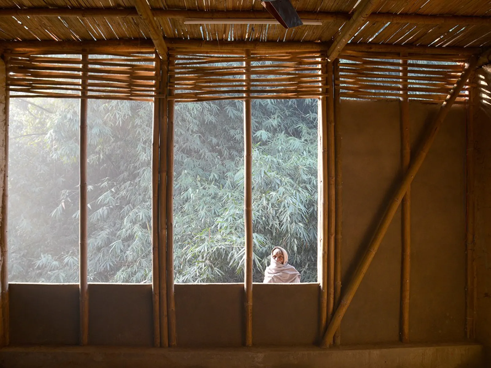 Innenansicht eines Gebäudes mit Wänden aus Lehm und Bambus, durch ein großes Fenster fällt Sonnenlicht auf eine Person, die draußen steht. Im Hintergrund sind Bäume zu sehen.