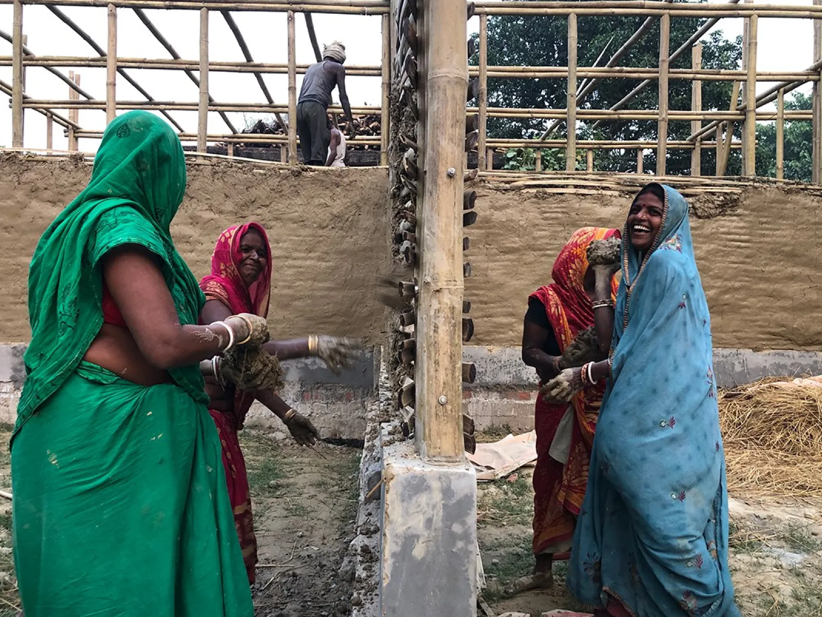Frauen in bunten Saris arbeiten gemeinsam an einem Lehmgebäude. Im Hintergrund ist eine Holzkonstruktion zu sehen. Sie lächeln und sind in den Bauprozess vertieft.