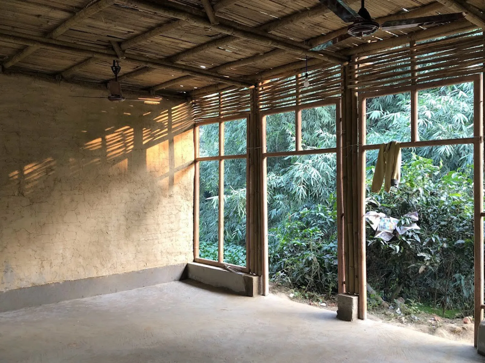 Innenansicht eines Schulgebäudes in Bahuarwa, Indien, mit Wänden aus natürlichen Materialien und großen Fenstern, die viel Tageslicht hereinlassen. Draußen ist üppige Vegetation zu sehen.
