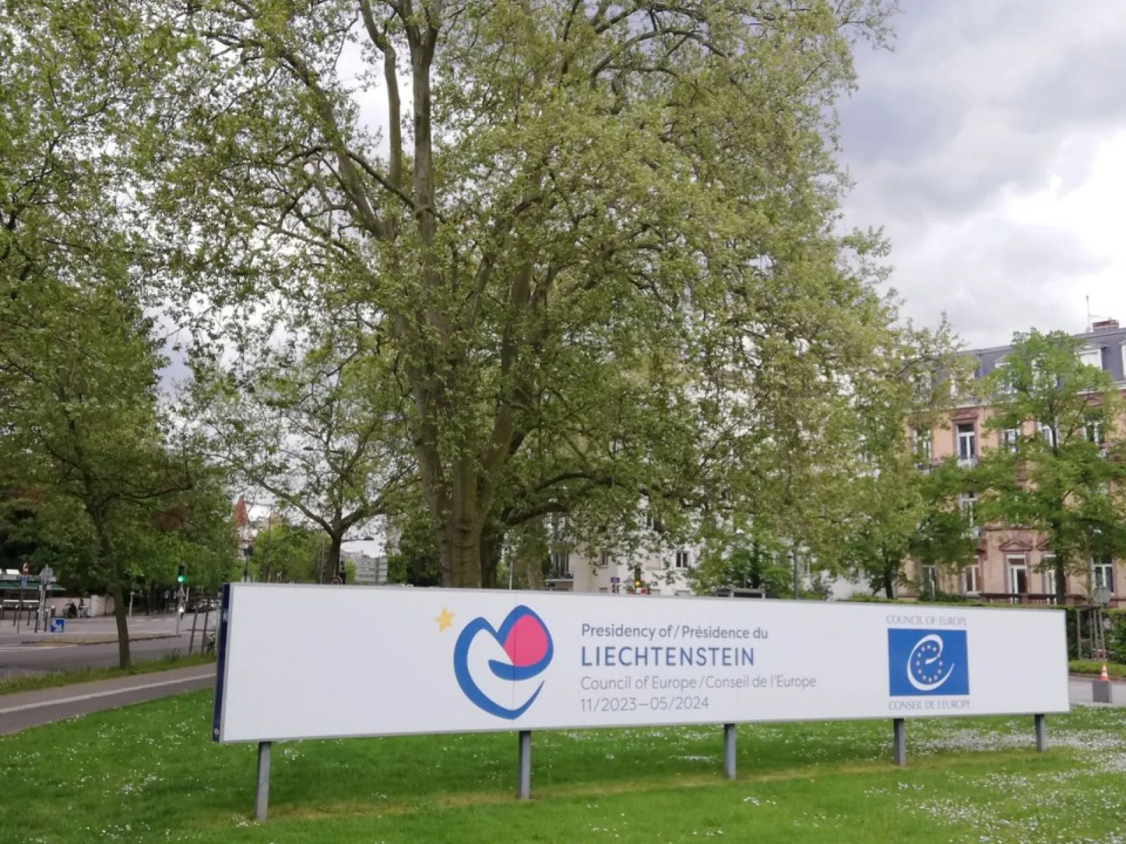 Ein großes Schild mit dem Logo des Fürstentums Liechtenstein und dem Europarat steht auf einer grünen Wiese, umgeben von Bäumen und Gebäuden im Hintergrund.