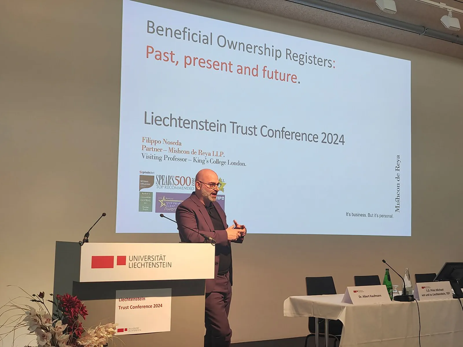 Ein Redner steht vor einem Podium mit dem Logo der Universität Liechtenstein und präsentiert bei der Liechtenstein Trust Conference 2024. Im Hintergrund ist eine Projektion mit dem Titel "Beneficial Ownership Registers: Past, present and future" zu sehen.