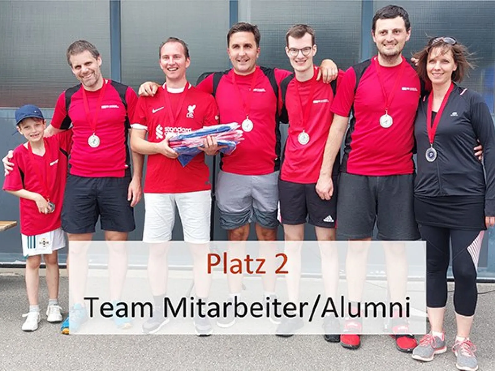 Gruppe von sechs Personen in roten Trikots mit Medaillen, die das Team "Mitarbeiter/Alumni" repräsentieren, posiert für ein Foto. Ein Kind steht links daneben. Ein Schild zeigt "Platz 2". Hintergrund: Gebäude mit Glasfront.