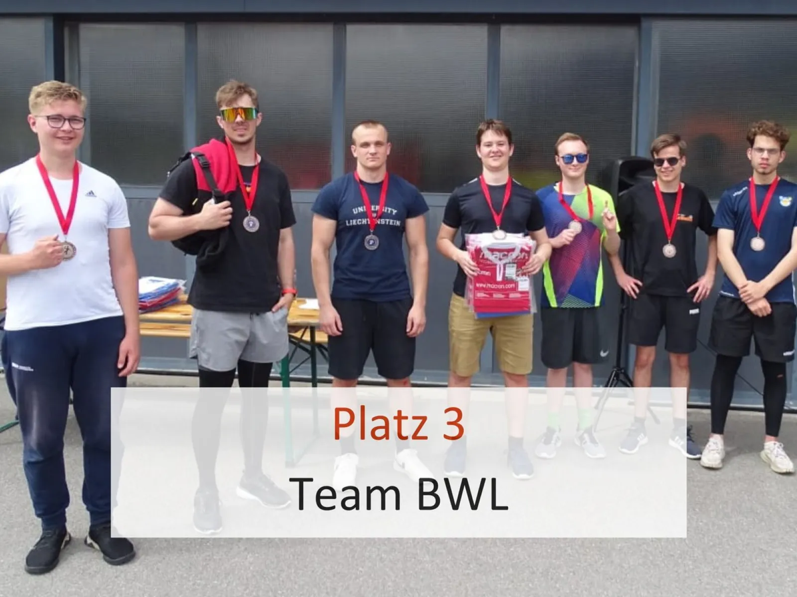 Eine Gruppe von sieben jungen Männern steht nebeneinander und trägt Medaillen um den Hals. Sie halten eine Urkunde und lächeln in die Kamera. Im Vordergrund steht der Text "Platz 3 Team BWL".