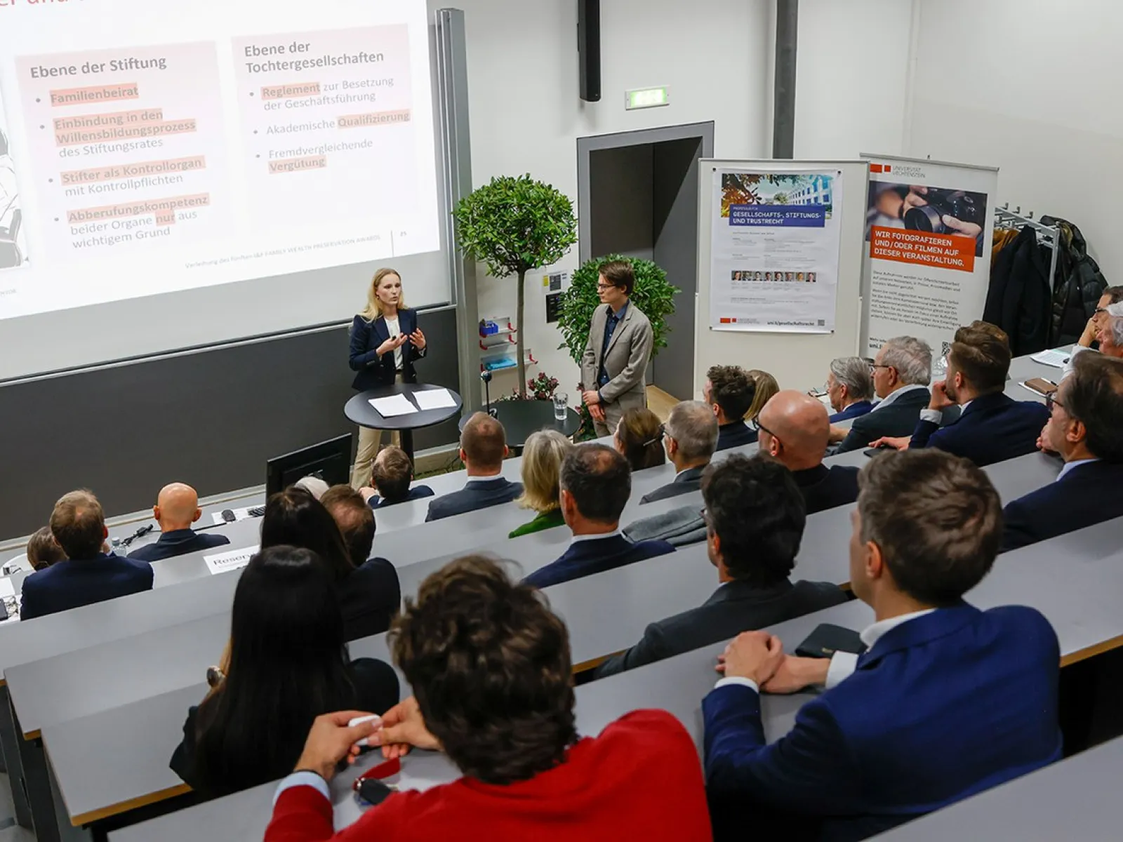 Ein Vortrag in einem Hörsaal der Universität Liechtenstein: Eine Person präsentiert vor einer Gruppe von Zuhörern, die in gestaffelten Sitzreihen sitzen. Im Hintergrund sind eine Leinwand mit Präsentationsfolien und ein Roll-up-Banner zu sehen.