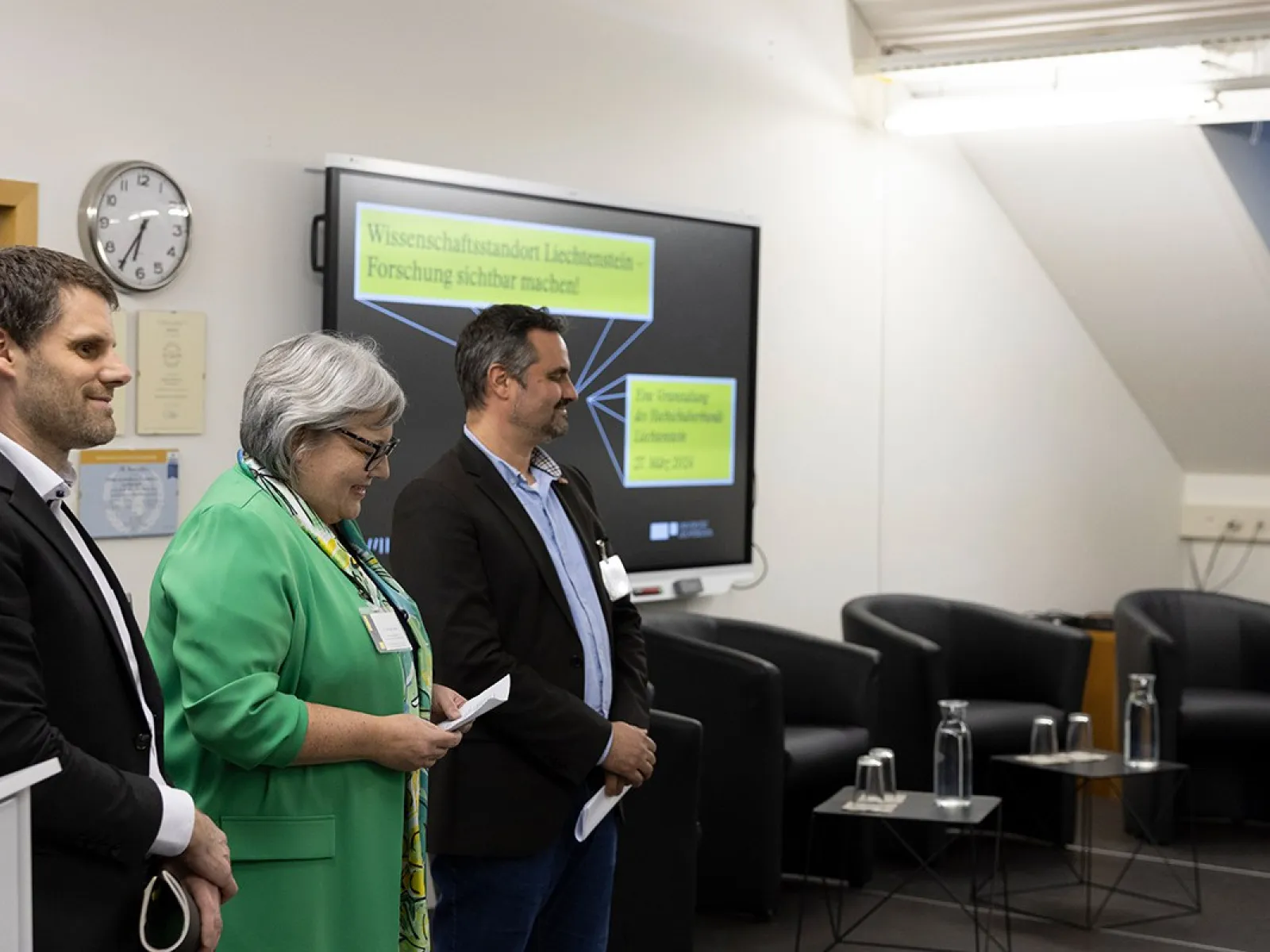 Drei Personen stehen nebeneinander in einem Konferenzraum. Im Hintergrund ist ein Bildschirm mit der Aufschrift "Wissenschaftsverbund Liechtenstein" zu sehen. Links im Bild ist ein Rednerpult, rechts sind leere Stühle und Tische. Die Personen tragen Namensschilder und lächeln.