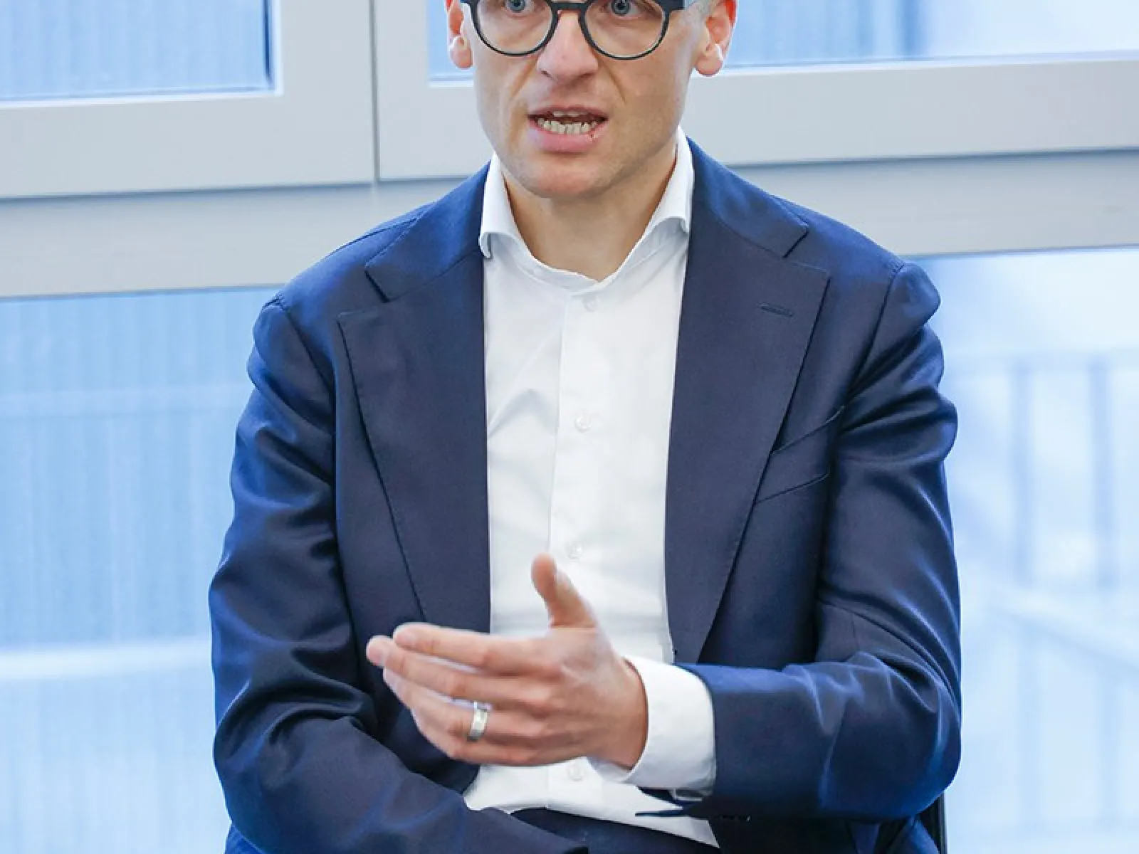 Ein Mann in einem Anzug spricht gestikulierend während einer Podiumsdiskussion in einem modernen Raum mit großen Fenstern.