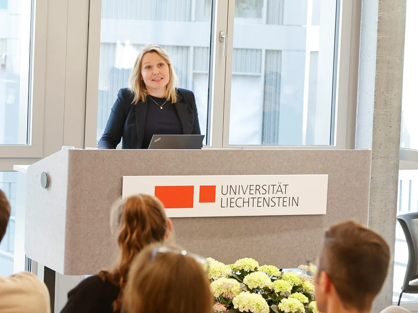 Eine Person steht an einem Rednerpult der Universität Liechtenstein und spricht zu einem Publikum. Im Hintergrund sind große Fenster zu sehen. Vor dem Pult sind Blumen arrangiert.