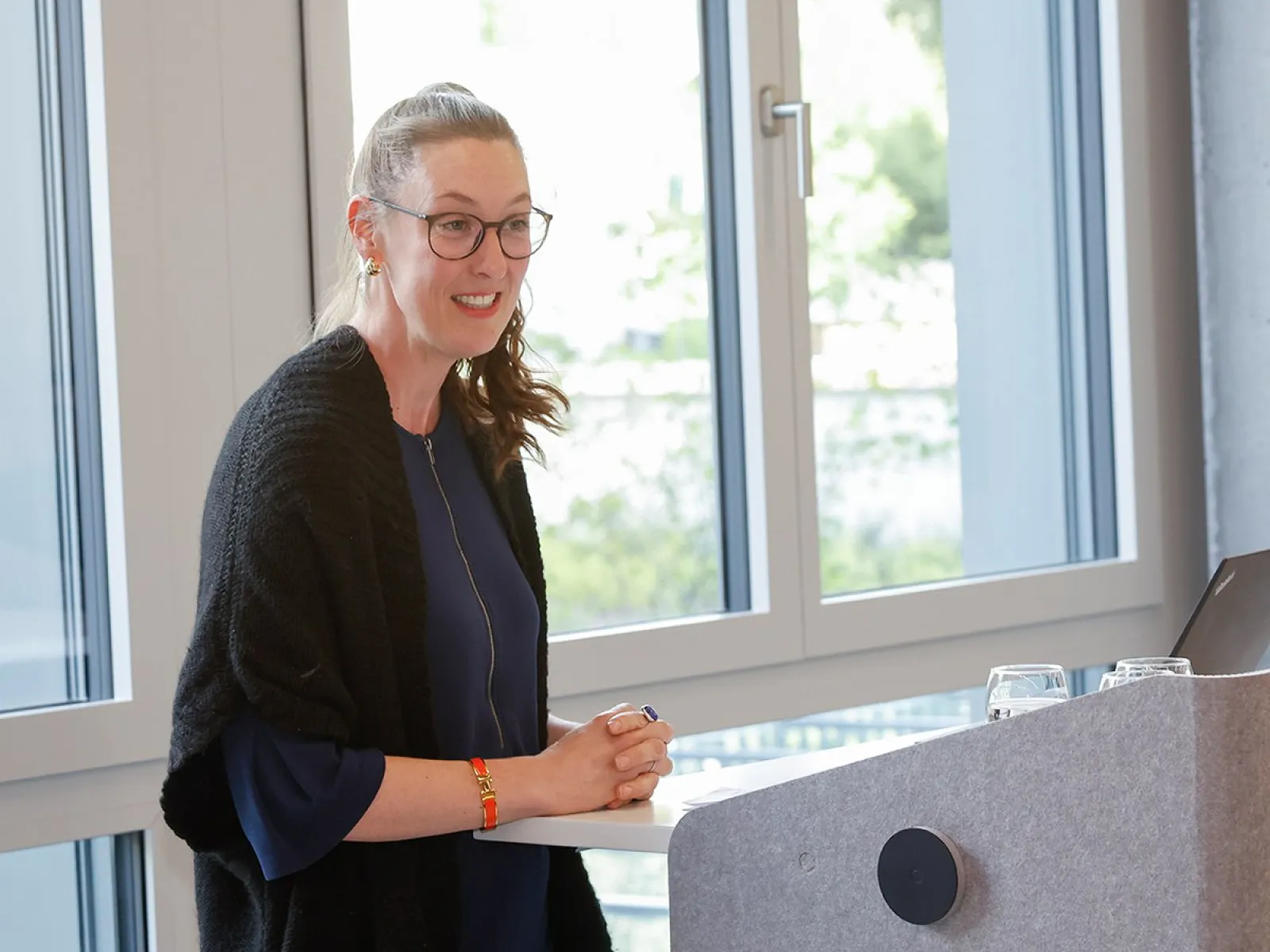 Eine Person steht lächelnd an einem Rednerpult in einem modernen Raum mit großen Fenstern. Sie trägt eine Brille und hält einen Stift in der Hand. Im Hintergrund sind Bäume zu sehen.