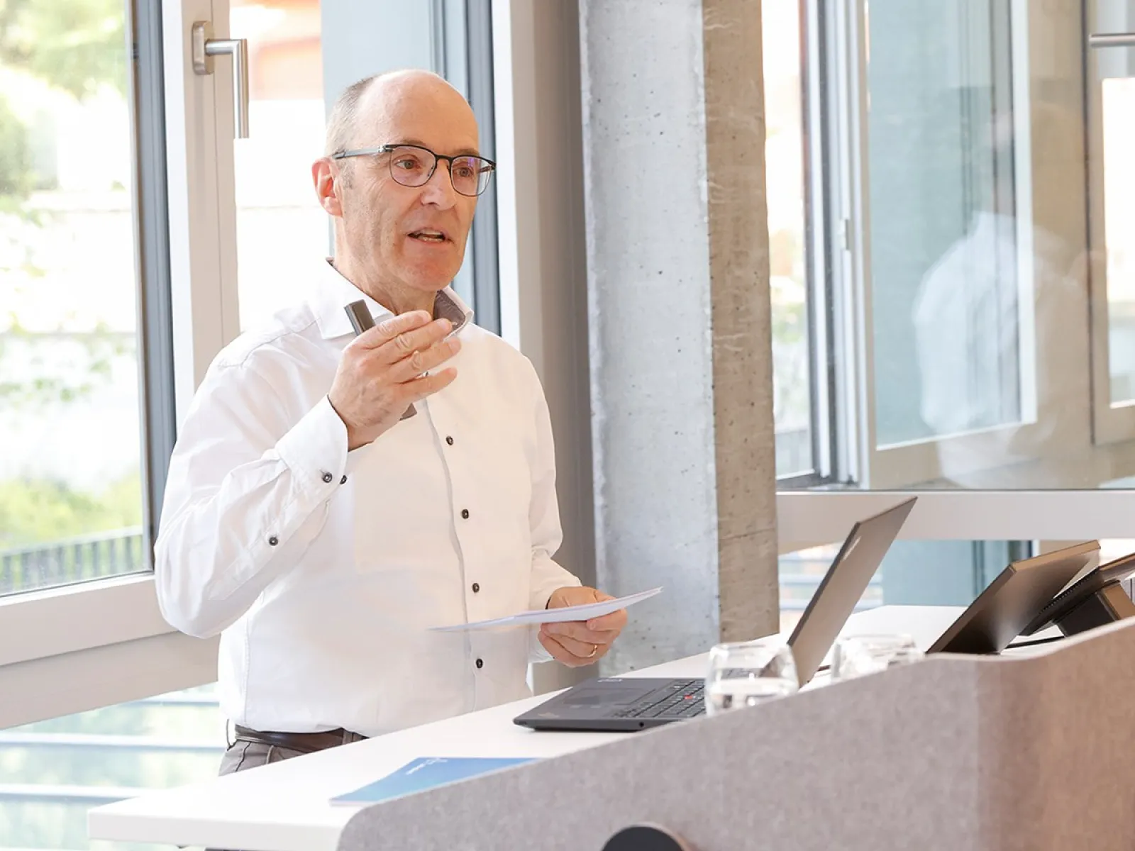 Ein Mann hält einen Vortrag in einem modernen Raum mit großen Fenstern. Er steht an einem Rednerpult mit Laptop und Notizen.