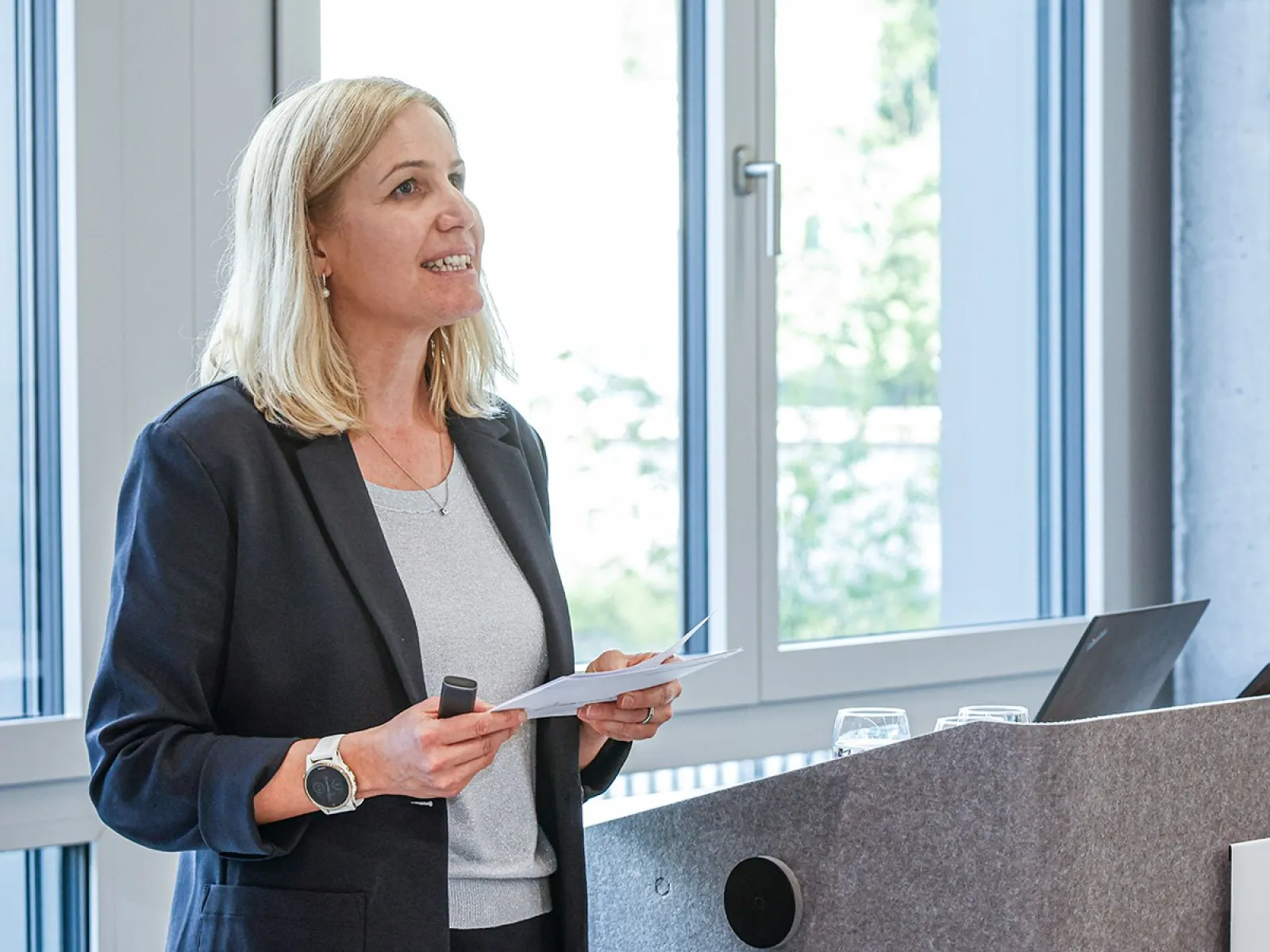 Eine Person hält einen Vortrag an einem Rednerpult in einem modernen Raum mit großen Fenstern. Sie trägt einen dunklen Blazer und hält ein Mikrofon sowie Notizen in der Hand.
