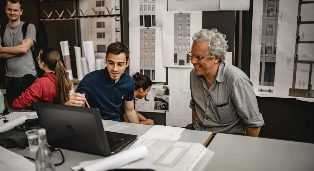 Dozent und Student sitzen lachend an einem Tisch und unterhalten sich über Architekturpläne 