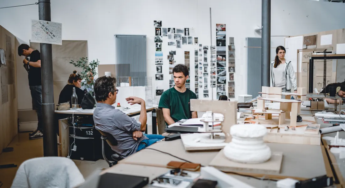 Dozent im Gespräch mit einem Architekturstudenten im offenen Atelier der Universität Liechtenstein, im Hintergrund weitere kreative Arbeitsprozesse.