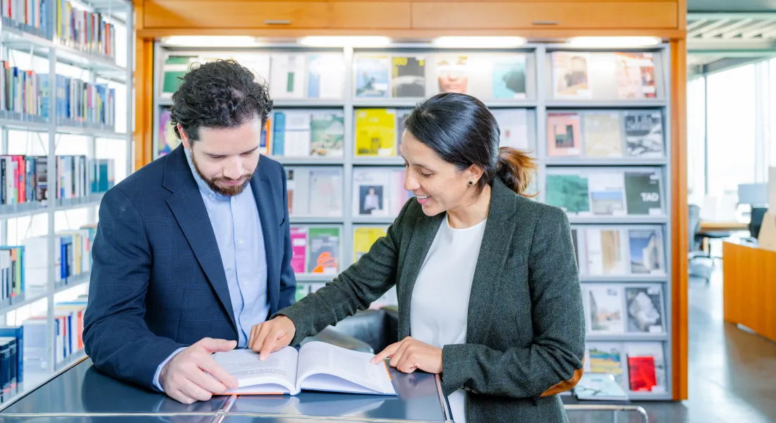 Zwei professionell gekleidete Personen in der Bibliothek der Universität Liechtenstein betrachten gemeinsam ein Buch und diskutieren dessen Inhalt, im Hintergrund Fachzeitschriftenregale.
