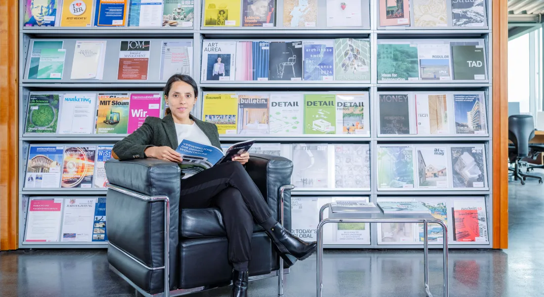 Frau sitzt entspannt auf einem schwarzen Ledersessel in der Bibliothek der Universität Liechtenstein und liest in einer Fachzeitschrift, im Hintergrund ein Regal mit bunten Magazincovern.