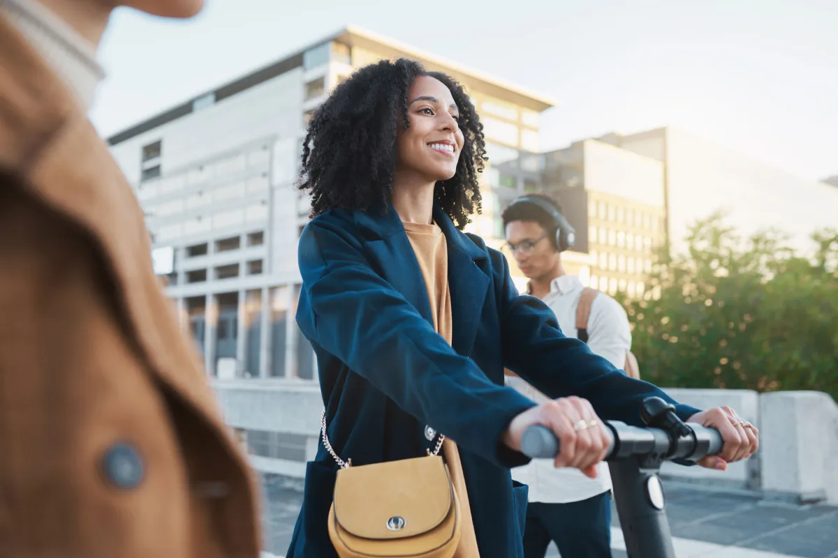 Eine junge Frau fährt auf einem E-Scooter durch eine städtische Umgebung auf dem Weg zur Universität. Die Gebäude im Hintergrund sind unscharf und deuten auf eine moderne Stadtlandschaft hin. Die Frau trägt keine Schutzkleidung und scheint sich sicher und zufrieden auf ihrem Weg zu befinden, wobei der E-Scooter als umweltfreundliche Transportlösung hervorsticht.
