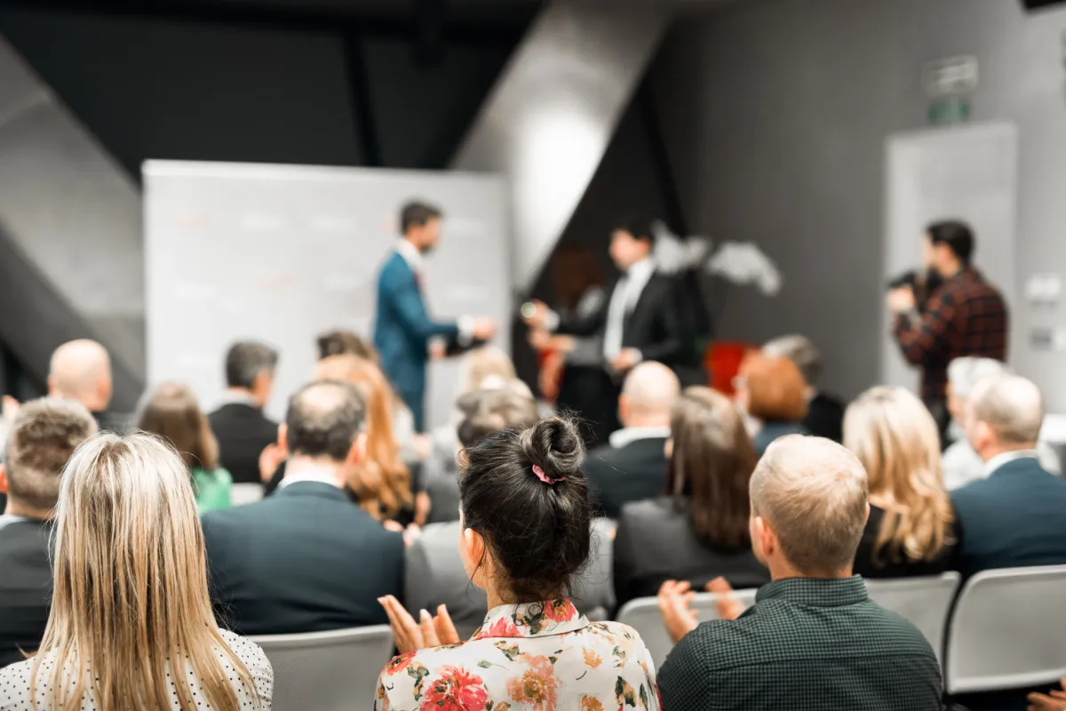 Ein Publikum sitzt in einem Konferenzraum und blickt nach vorne, wo zwei unscharf dargestellte Männer auf einer Bühne stehen. Einer der Männer überreicht dem anderen etwas, während die Zuschauer im Vordergrund applaudieren.