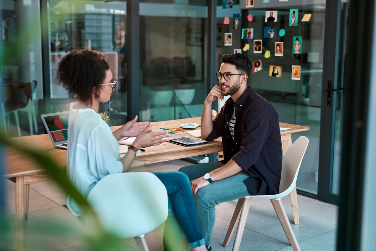Eine Frau und ein Mann sitzen sich in einem modernen Büro gegenüber. Die Frau spricht gestikulierend, während der Mann aufmerksam zuhört. Auf dem Tisch liegen ein Laptop und Büromaterialien. Im Hintergrund ist eine Glaswand mit bunten Haftnotizen und Fotos zu sehen.
