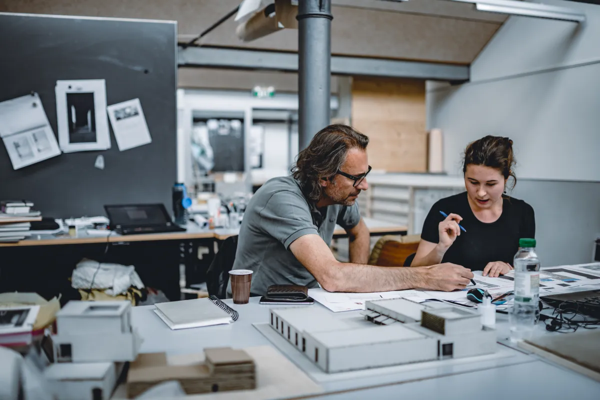 Dozent unterstützt eine Studentin bei der Projektarbeit im Architekturatelier der Universität Liechtenstein, umgeben von Modellen und Unterlagen.